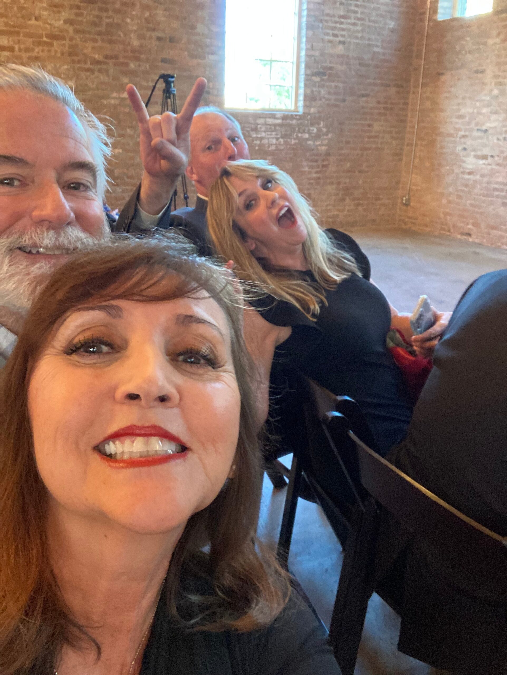 Close-up group selfie in a brick-walled room: smiling people, with a man in back making a rock-on hand gesture behind a woman in the foreground.