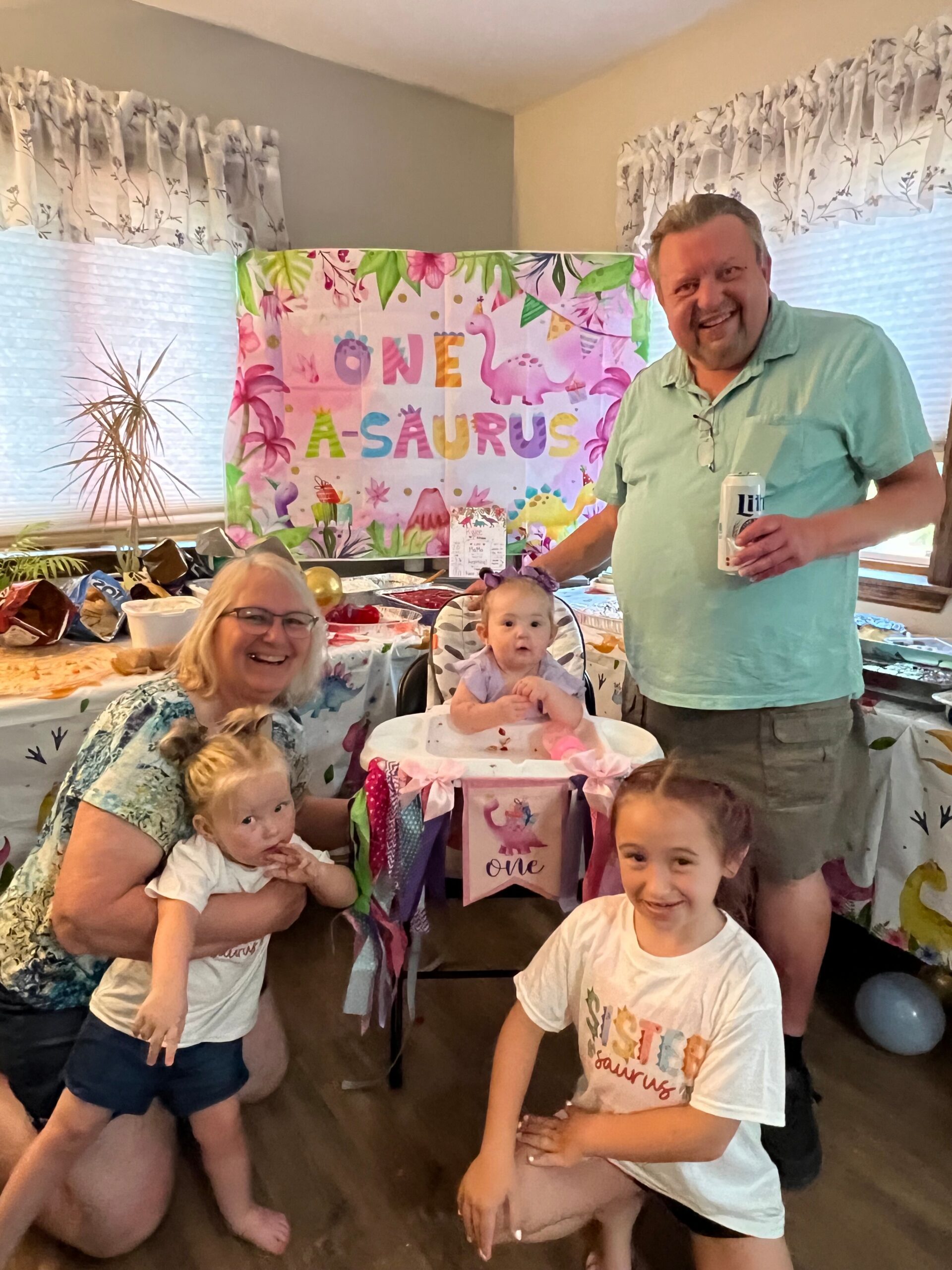 Family birthday party for a one-year-old with a colorful dinosaur banner that reads 'One- A-saurus' and smiling adults nearby.