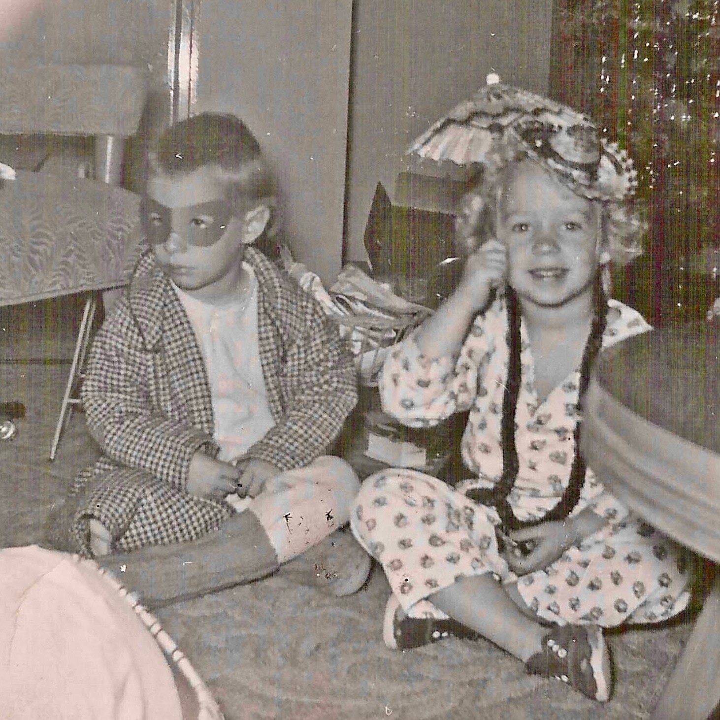 Two children in costumes sit on the floor indoors; the girl on the right wears a floral pajama-style outfit with braids and a feathered headpiece, smiling. The boy on the left wears a mask and a checkered coat over a light shirt.] } , {