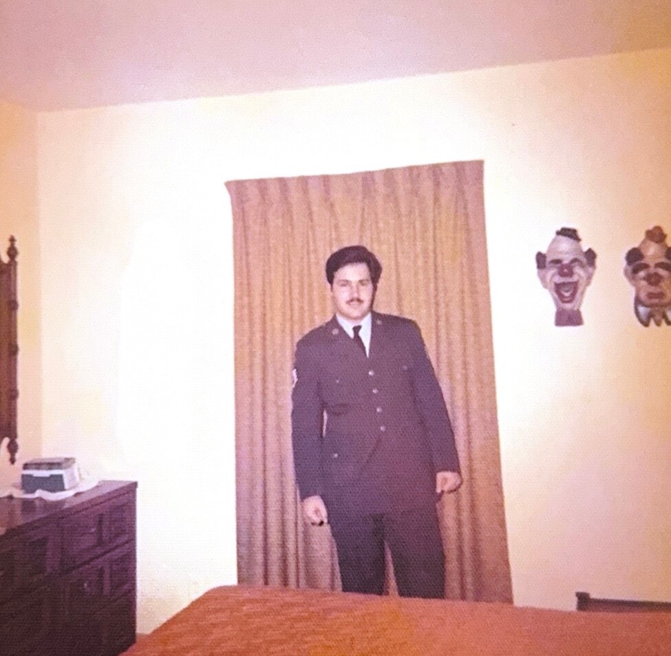 Man in a dark law enforcement uniform standing in a living room with a curtain backdrop and decorative wall masks to the right.