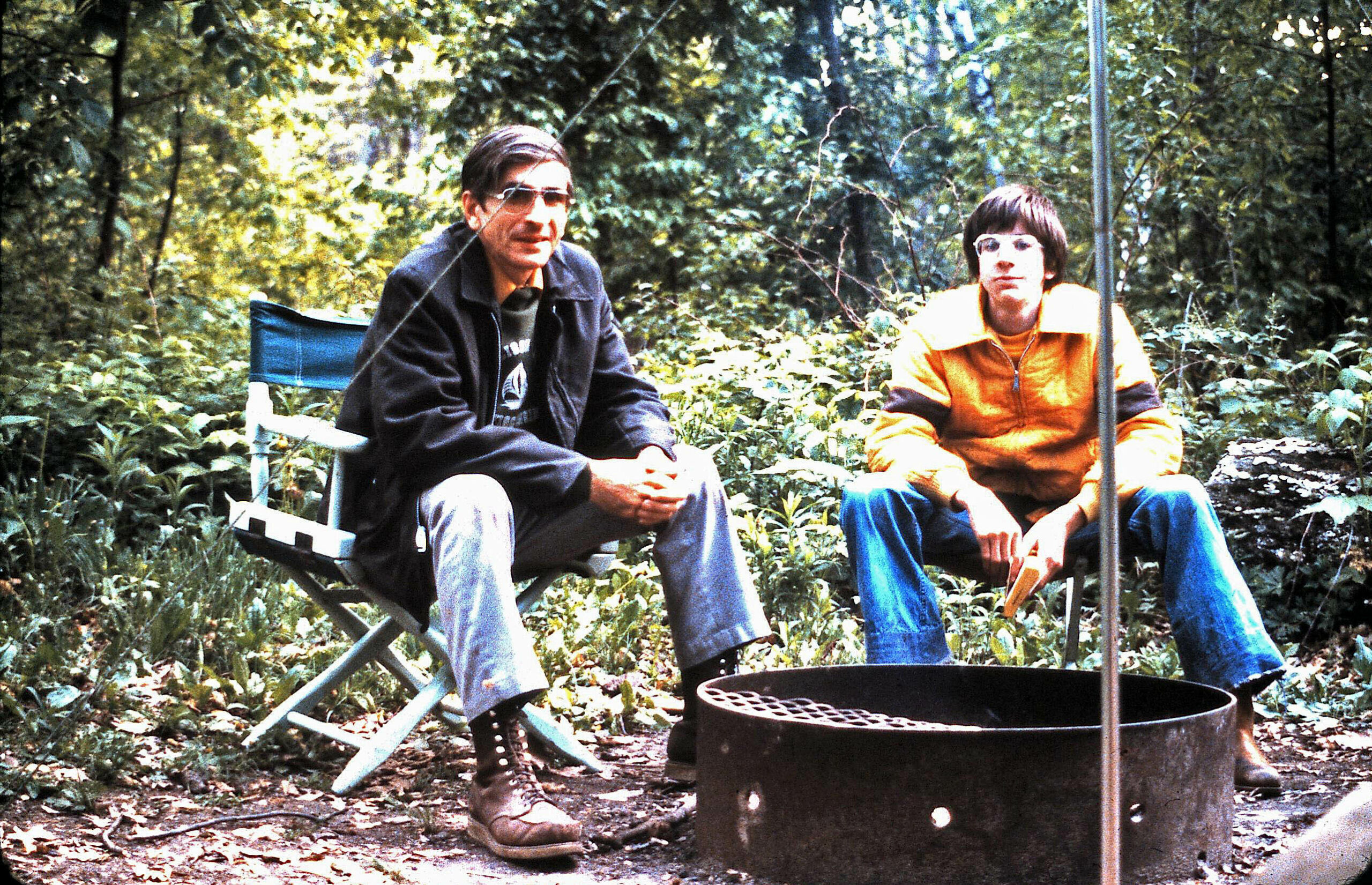 Two men sit on camping chairs around a metal fire pit in a wooded campsite.