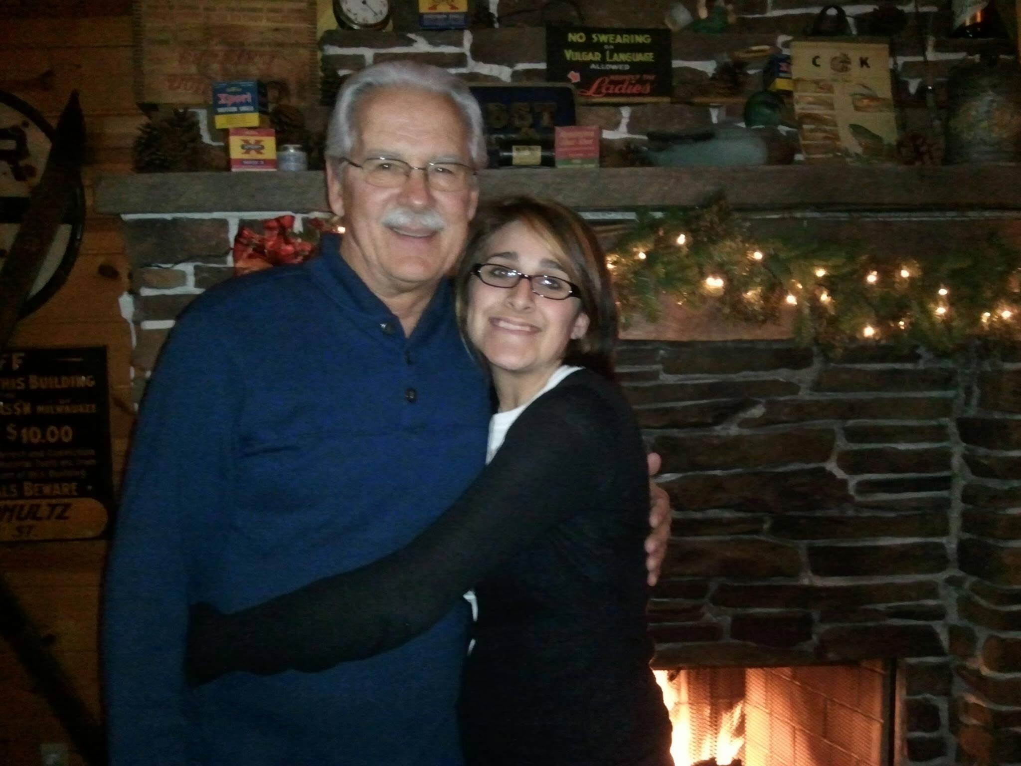 Smiling older man with glasses and a white mustache standing beside a younger woman, arms around each other in a cozy brick fireplace setting with string lights.