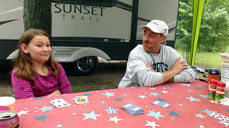 Girl in a pink shirt sits at a red star-patterned table, playing cards with a man in a gray hoodie and white cap outdoors beside a camper labeled 'Sunset Trail'.],