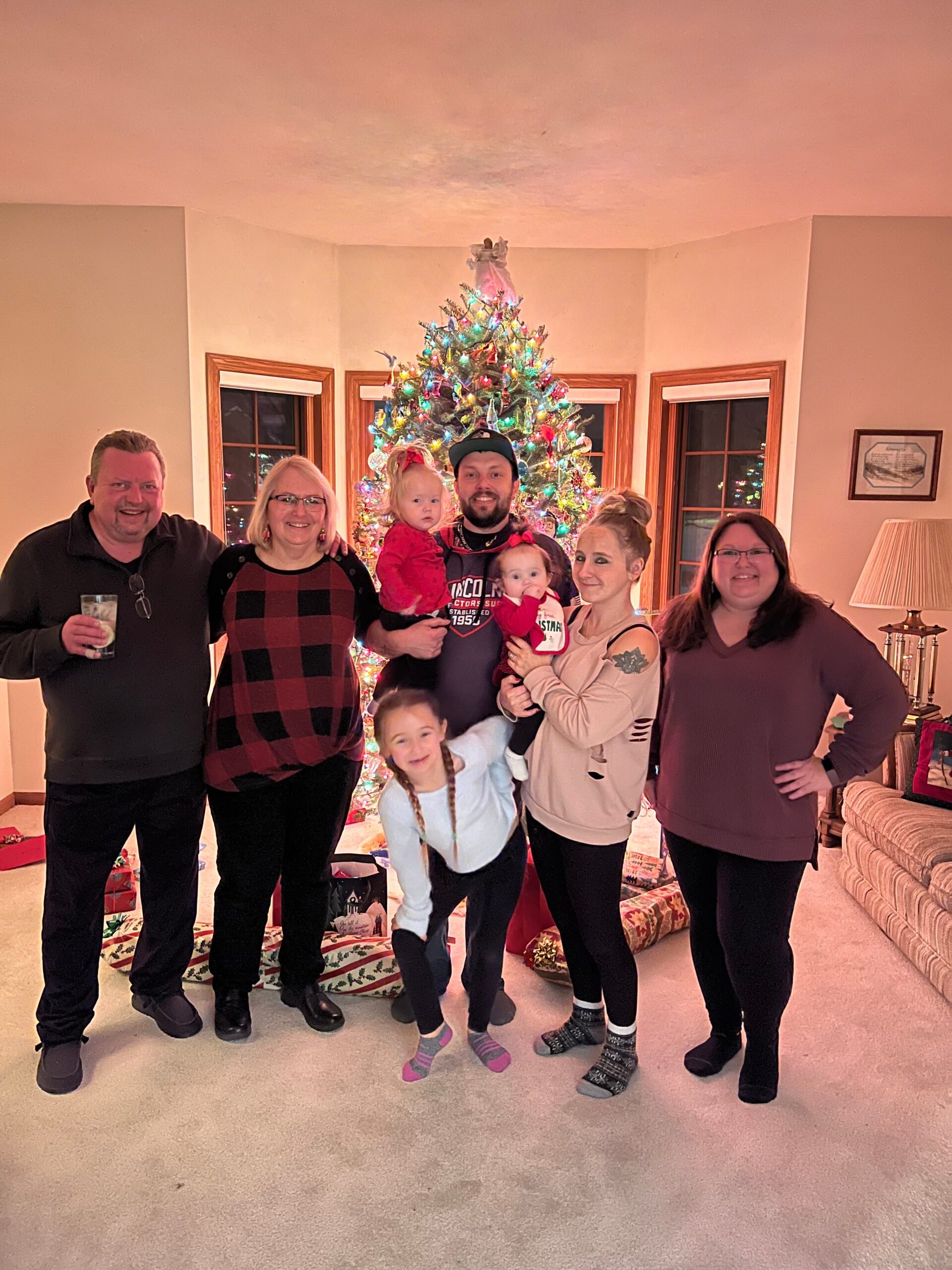 Group of seven people (including two young children) posing in a living room in front of a decorated Christmas tree with gifts underneath and warm lighting in the background.