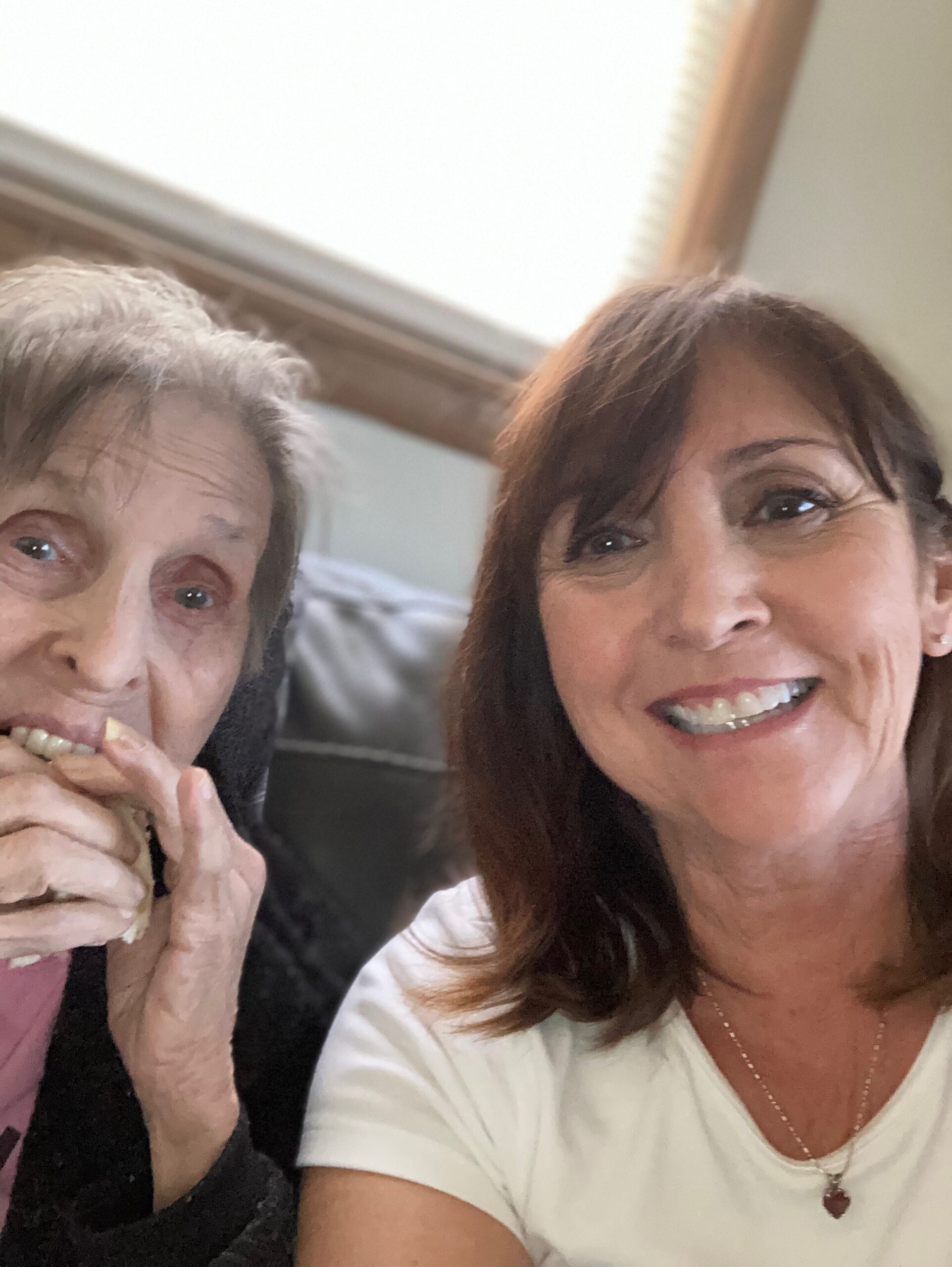 Two women posing for a selfie indoors; an older woman on the left with gray hair and a playful expression, and a smiling younger woman on the right with brown hair.
