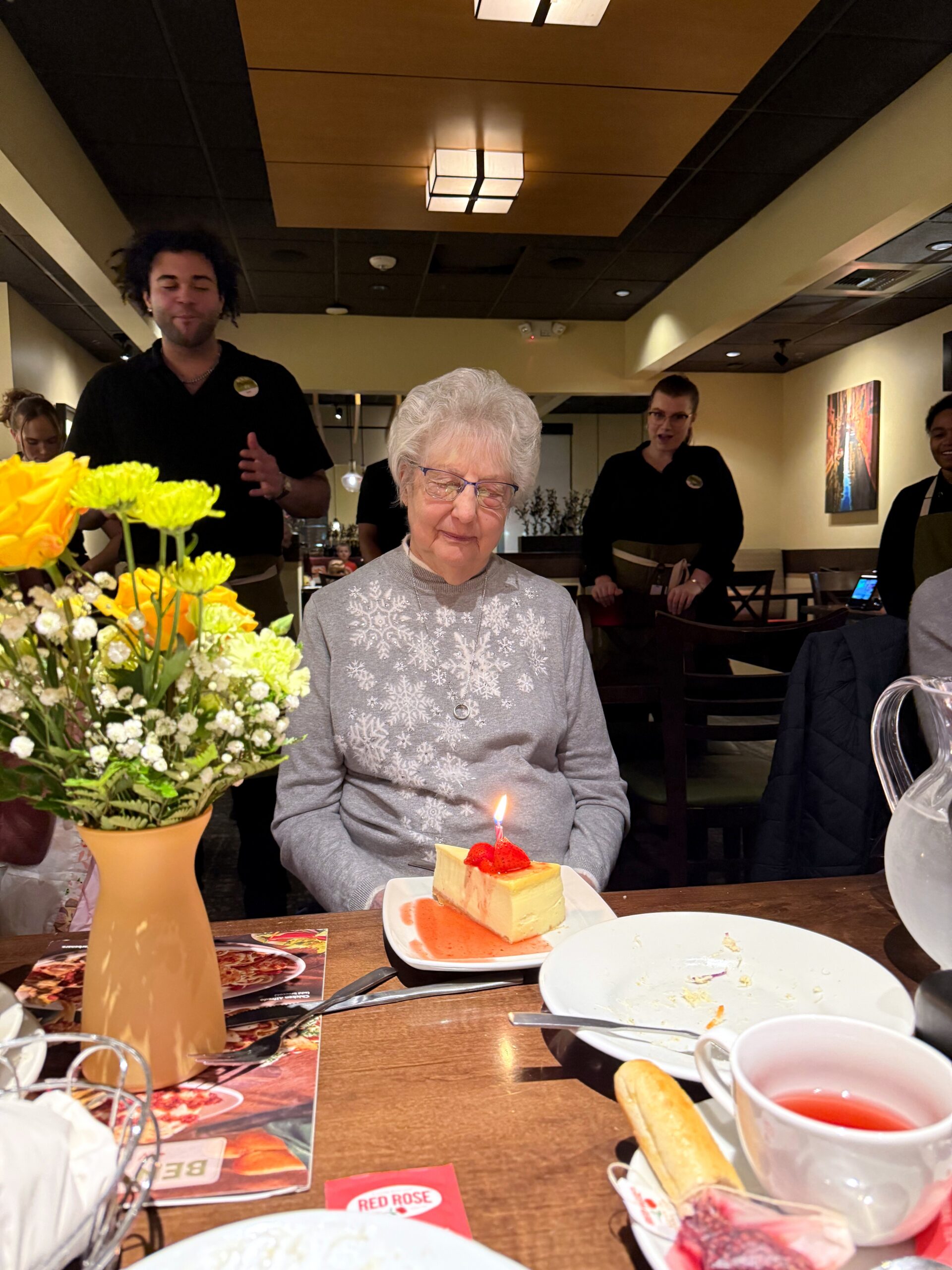 Older woman with silver hair at a restaurant table, looking down at a small cake with a lit candle, flowers in a vase nearby.