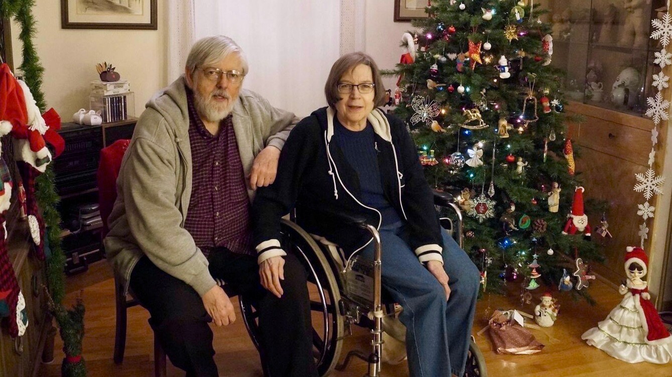 An elderly man and woman sit together in a living room beside a decorated Christmas tree.
