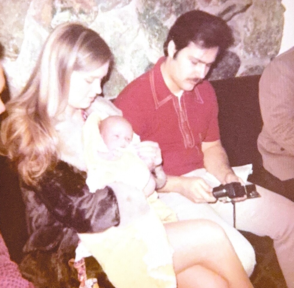 Young woman cradles a newborn while a man with a mustache sits beside her, holding a camera on his lap.