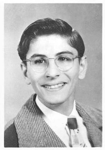 Black-and-white portrait of a young man with short dark hair and glasses, wearing a suit jacket over a white shirt with a striped tie, and smiling.