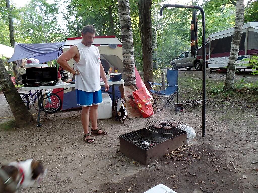 Man in blue shorts and sandals stands beside a charcoal grill cooking at a forest campsite with tents and camping gear nearby.