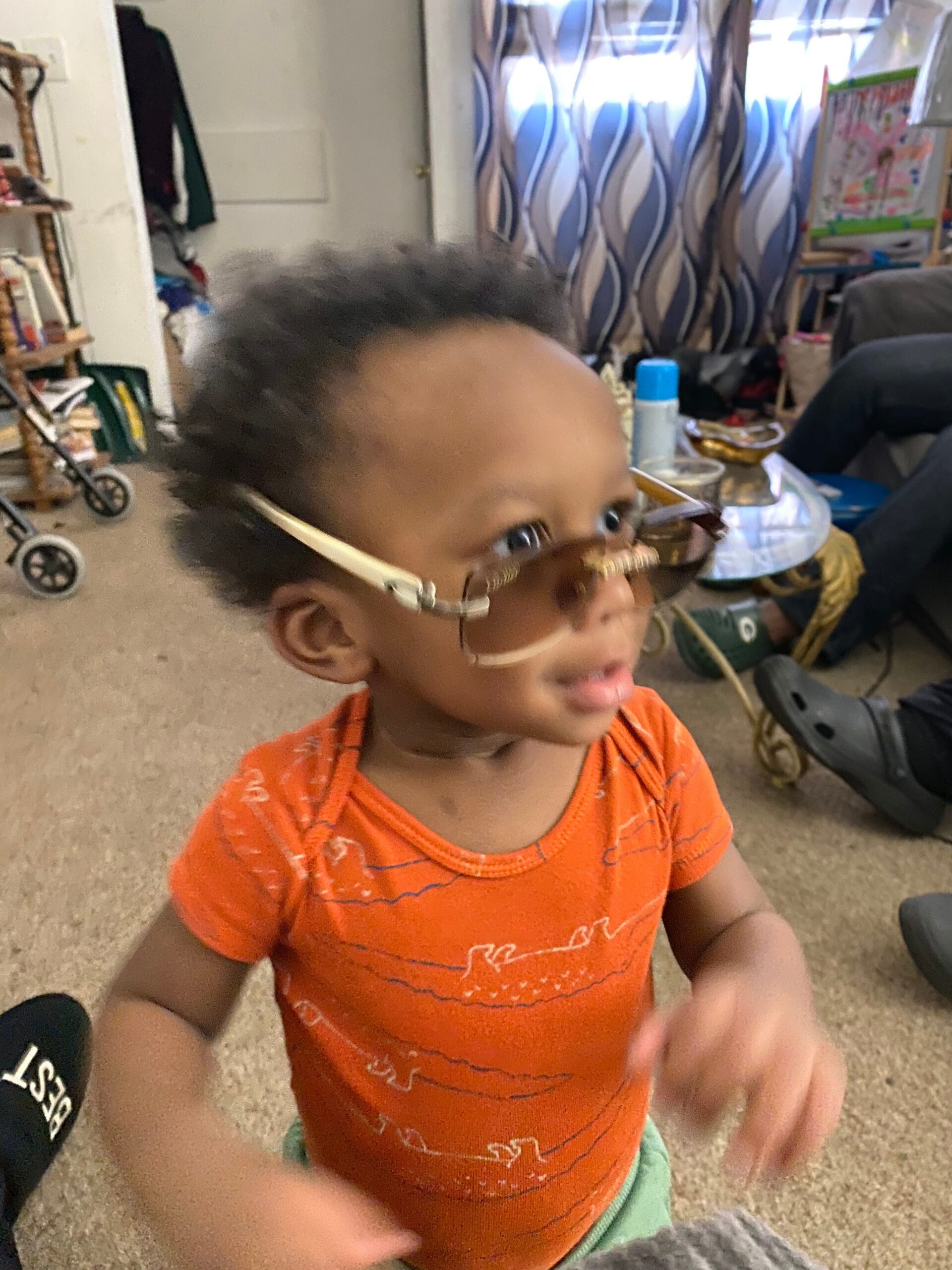 Young child with short curly hair wearing an orange shirt and clear glasses in a cluttered indoor room.