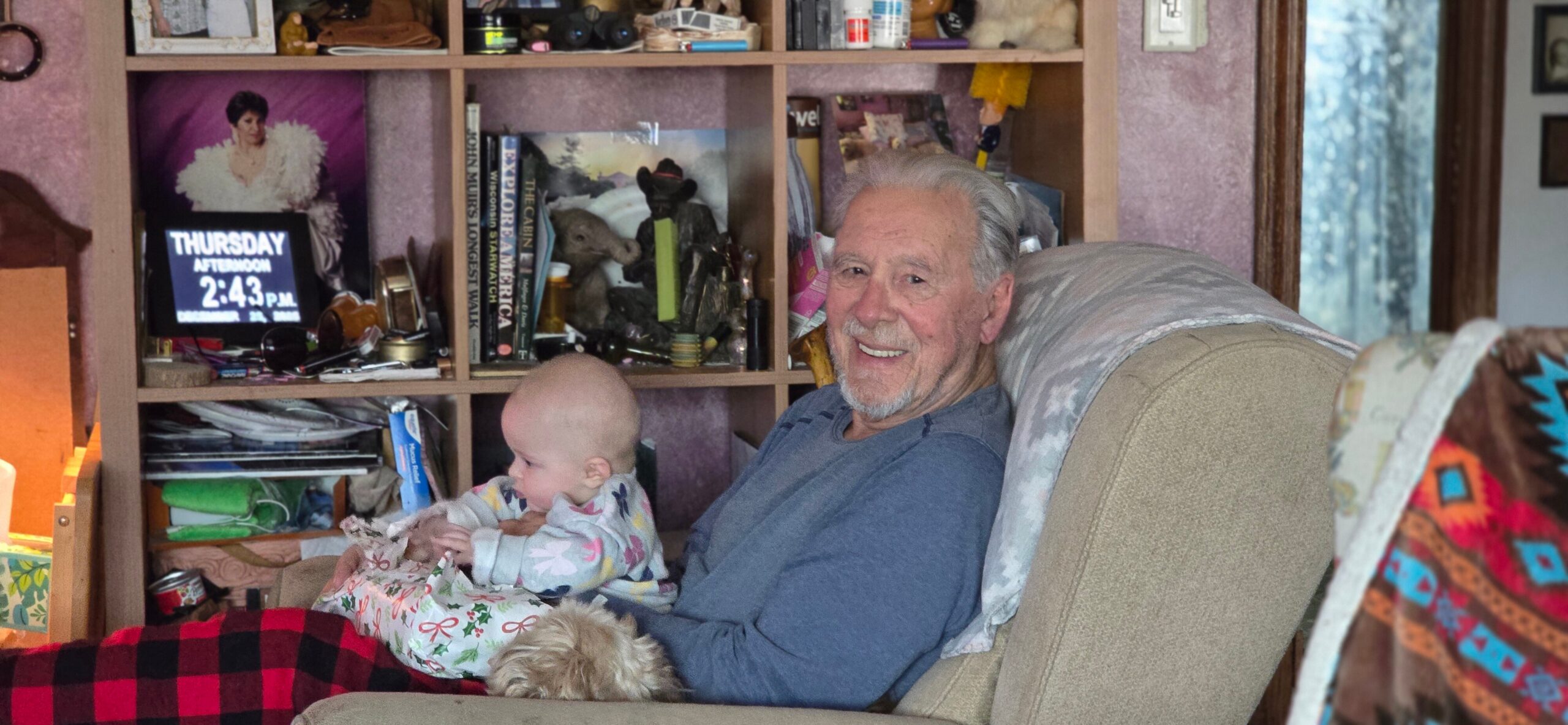 Older man in a blue shirt sits in a chair, smiling at a baby on his lap in a cozy, cluttered living room with a shelf of books behind them in view.