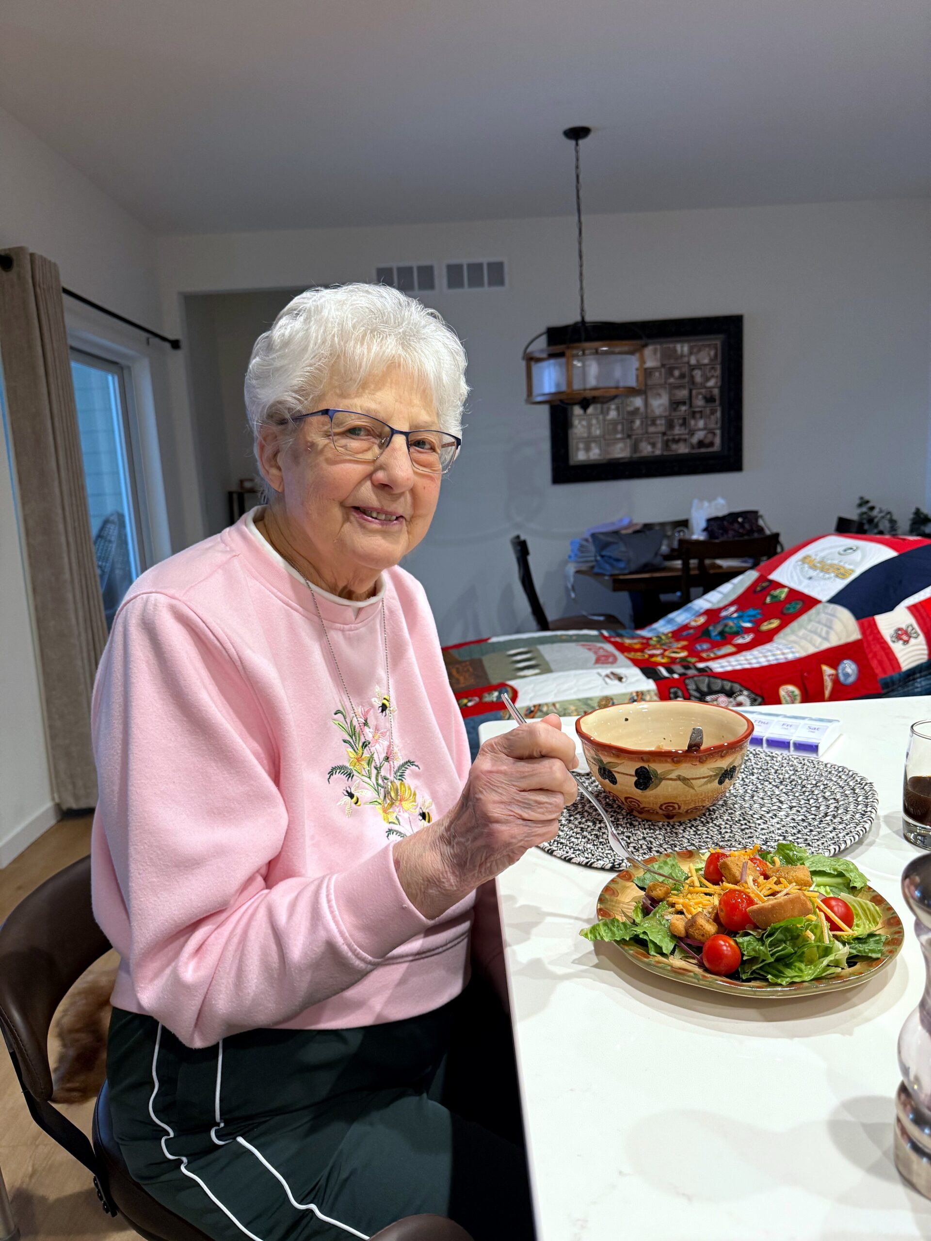 Smiling elderly woman with white hair and glasses sits at a dining table, eating a salad.