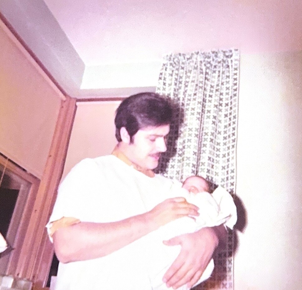 A man with dark hair and a mustache cradling a newborn baby wrapped in white, indoors near a patterned curtain.