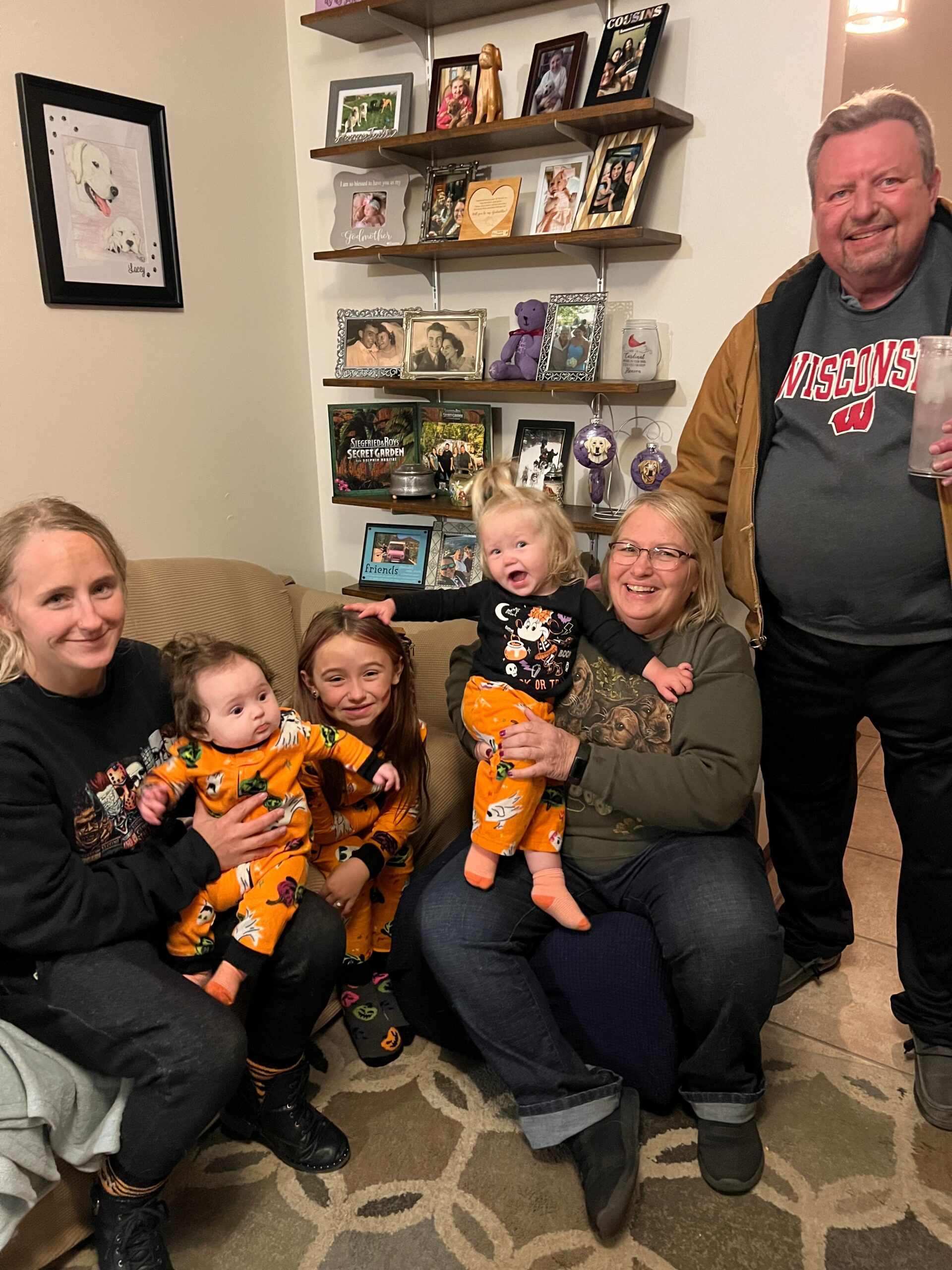 Family gathered on a couch, kids in Halloween outfits posing with smiling adults in a cozy living room with photo shelves behind them.