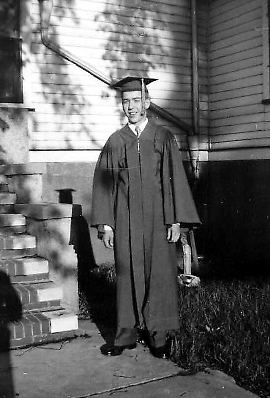 Graduate in cap and gown standing on a porch, holding a diploma.