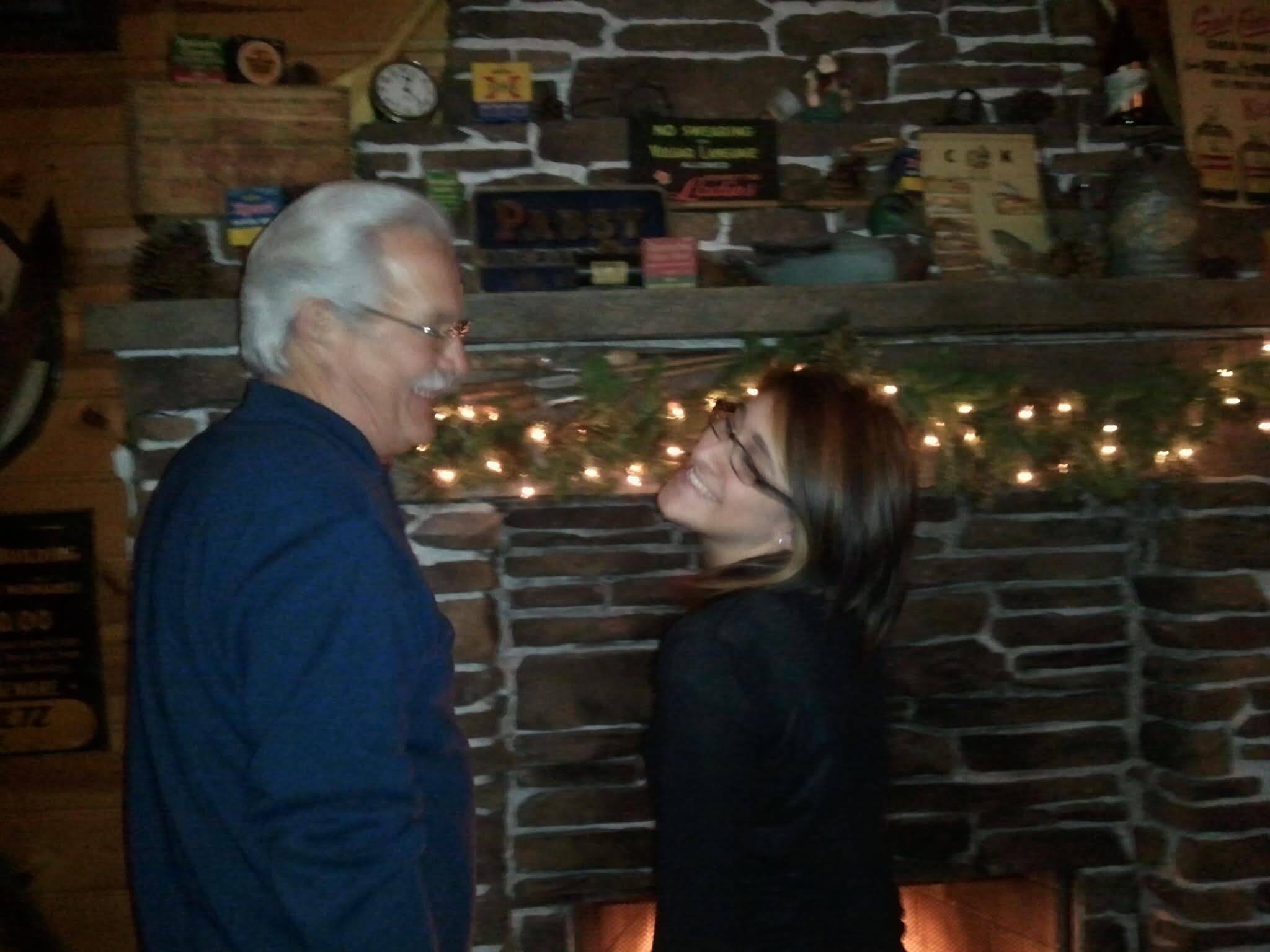 Two people smiling and facing each other indoors, standing in front of a stone fireplace with string lights and rustic decorations in the background.