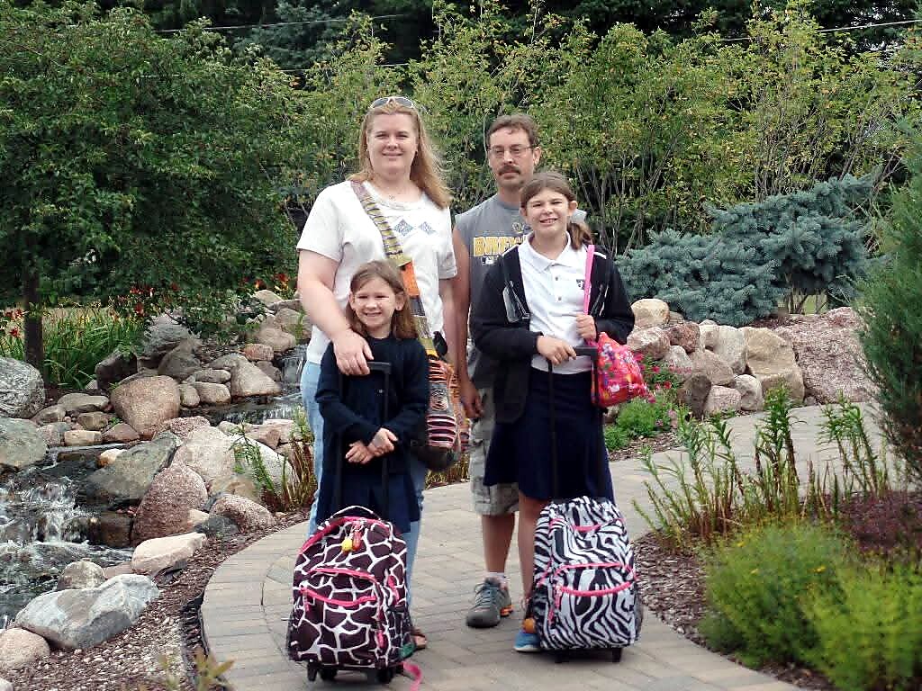 Family of four posing on a garden path with rolling suitcases and backpacks, likely ready for travel.