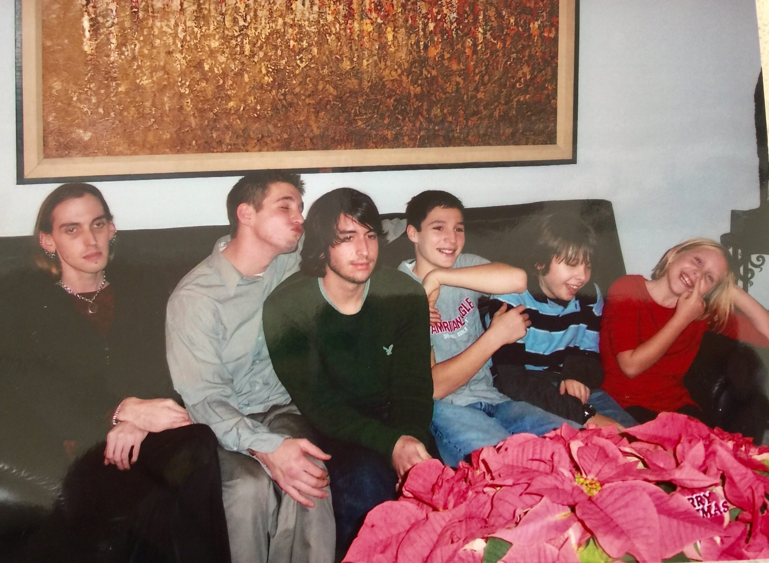 A group of seven friends sitting close together on a couch, posing for a casual photo with a large abstract painting on the wall behind them and pink flowers in the foreground.