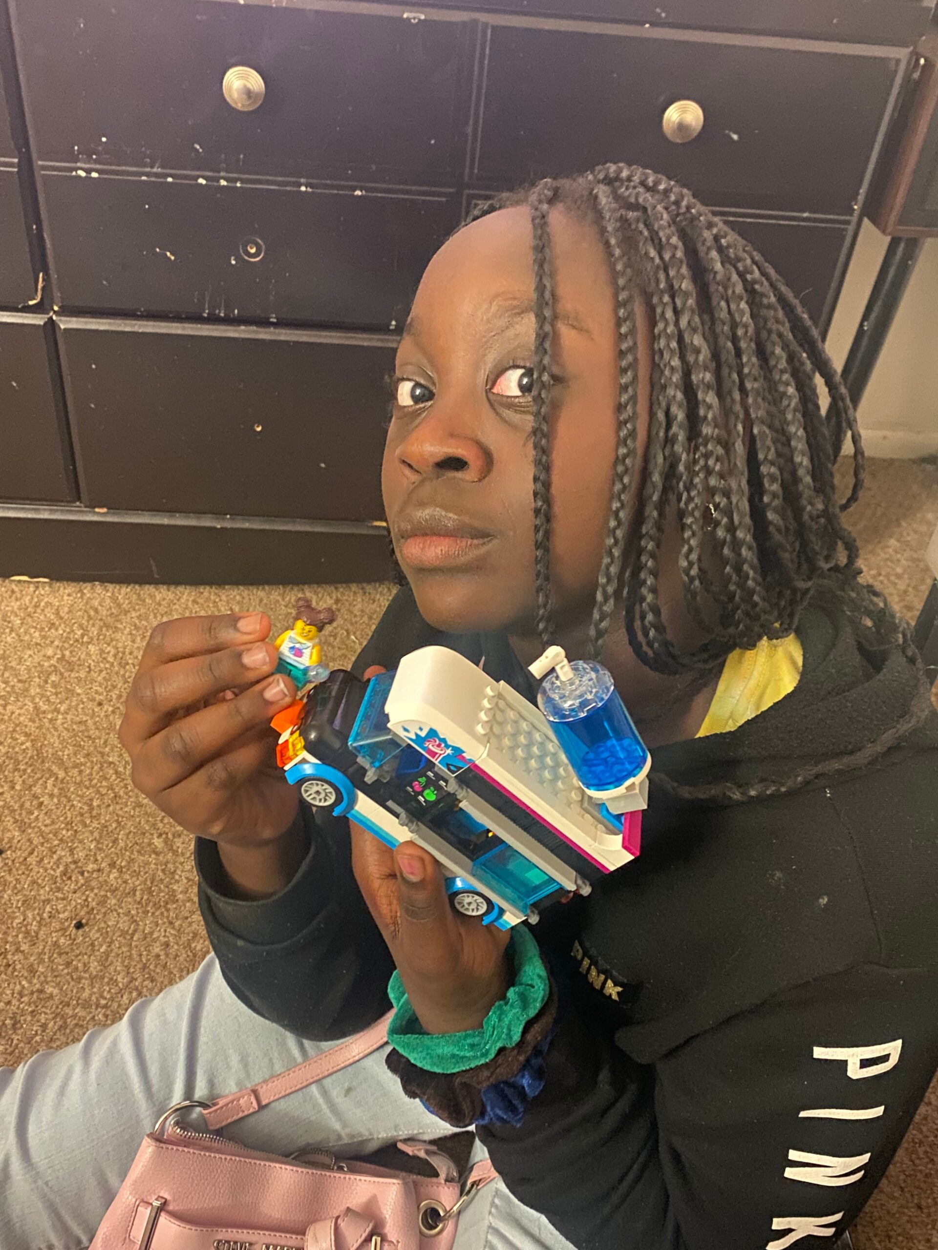 Young child with braided hair sits on the floor, playing with a blue-and-white LEGO vehicle and mini figures, with a dark dresser in the background.