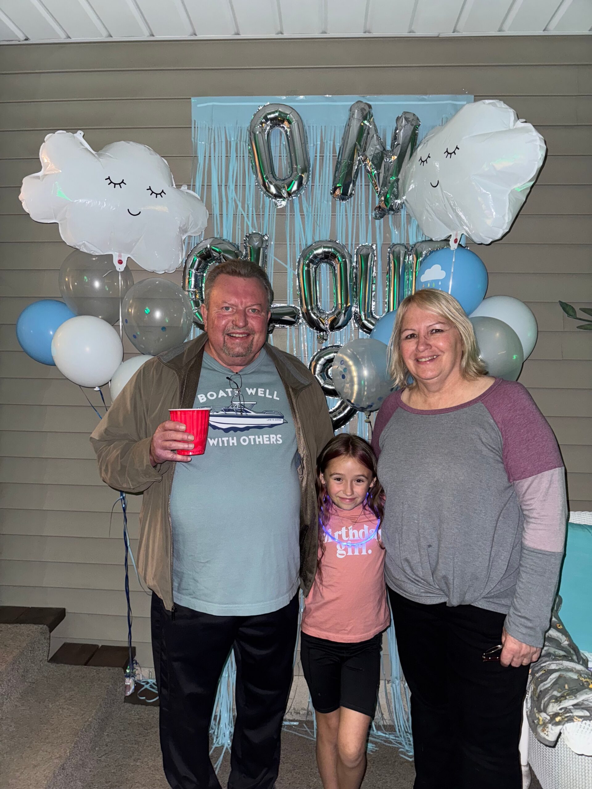 Family of three posing at a birthday party with blue/white balloons and paper cloud decorations behind them; man holds a red cup.