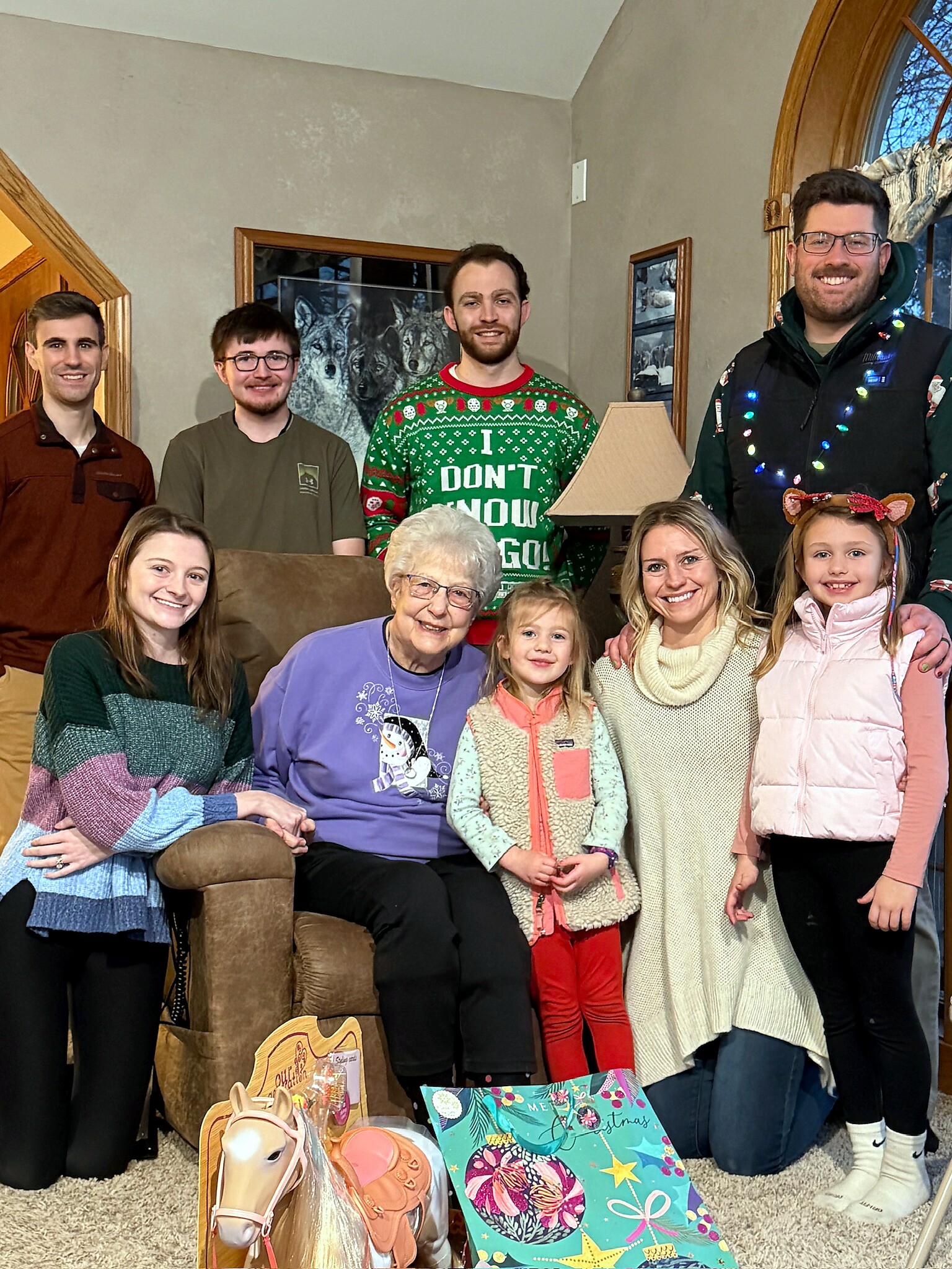 Group of eight people posing for a holiday photo in a living room with gift bags and a toy horse on the carpet.