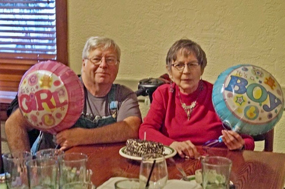 Two seniors sit at a dining table with a chocolate cake, holding pink 'It's a GIRL' and blue 'It's a BOY' balloons.