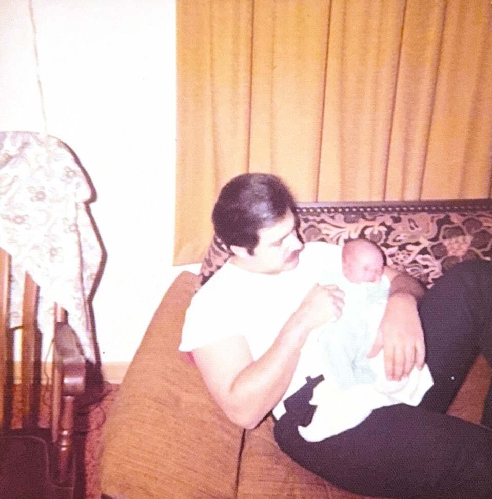Man sitting on a patterned couch cradling a newborn baby wrapped in a light blanket in a living room with yellow curtains on the window side.