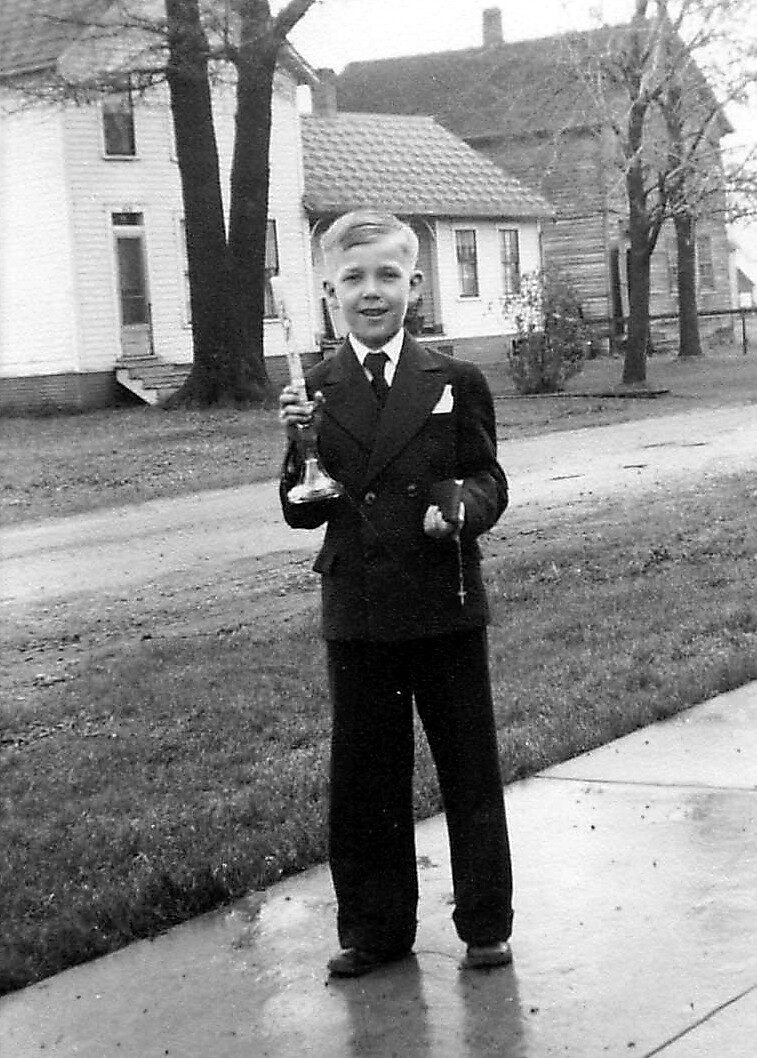 Boy in a formal suit standing on a wet sidewalk outside houses, holding a candle in one hand and a small item in the other.