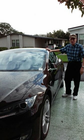 Older man wearing a cap and plaid shirt stands next to a shiny dark sedan in a residential driveway, his hand resting on the car body.