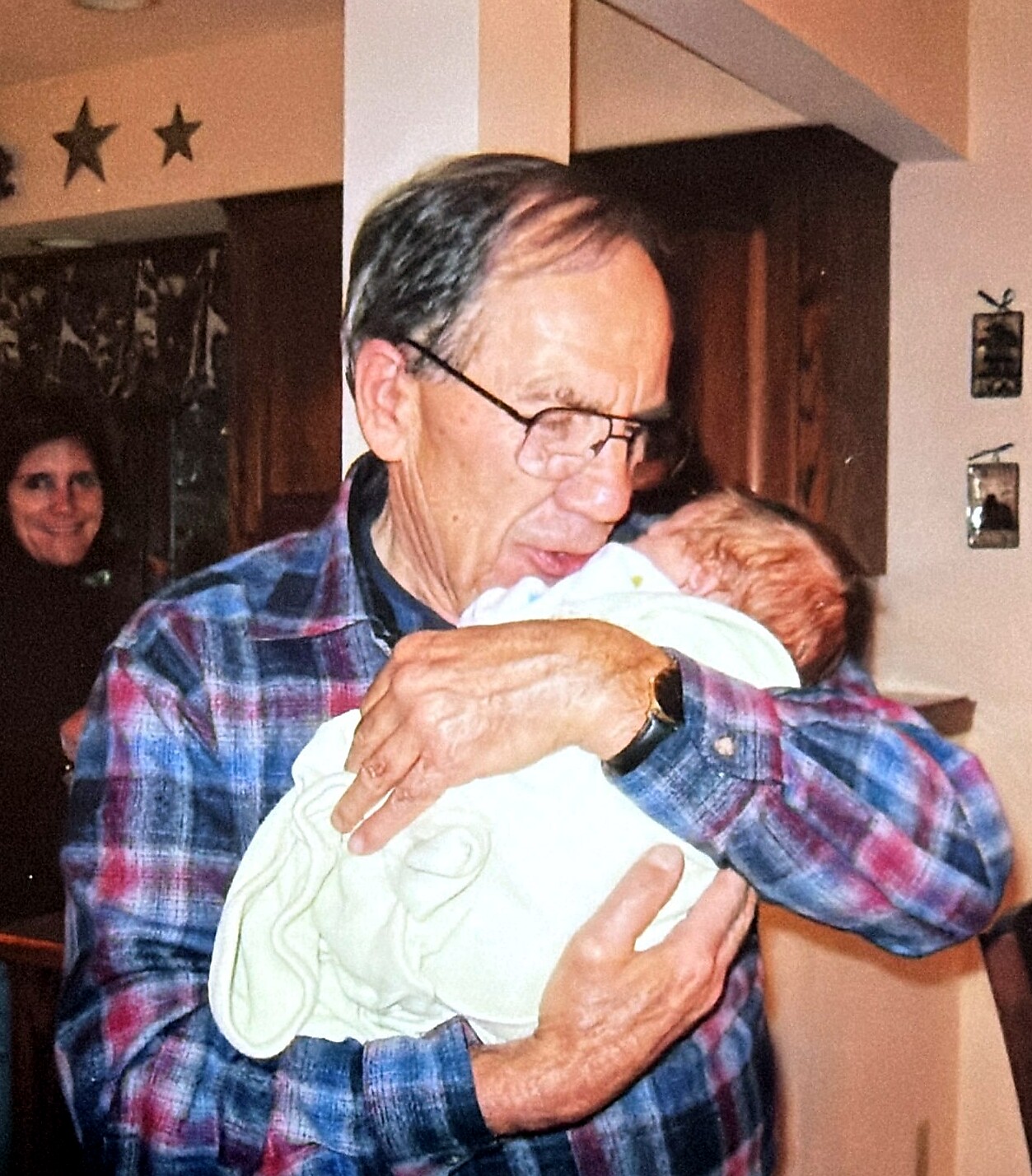 Older man in a plaid shirt hugging a baby wrapped in a light blanket indoors, with a smiling woman in the background.