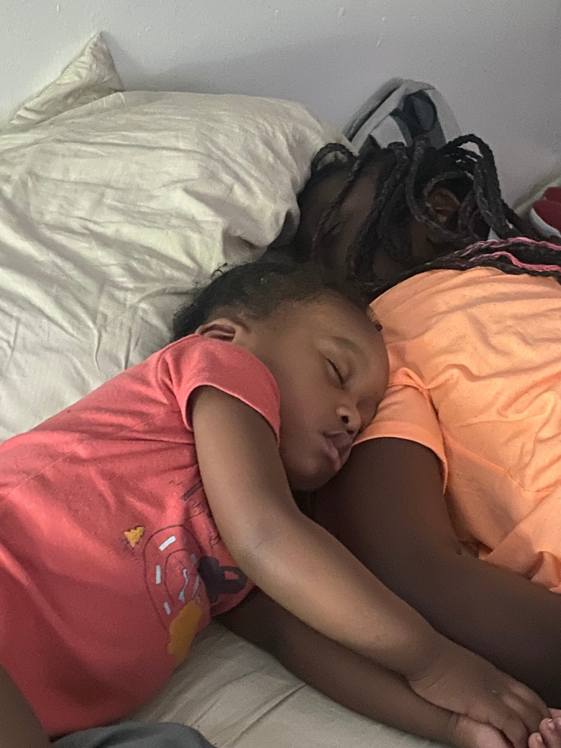 Two children asleep on a bed, cuddled together with white pillows behind them.