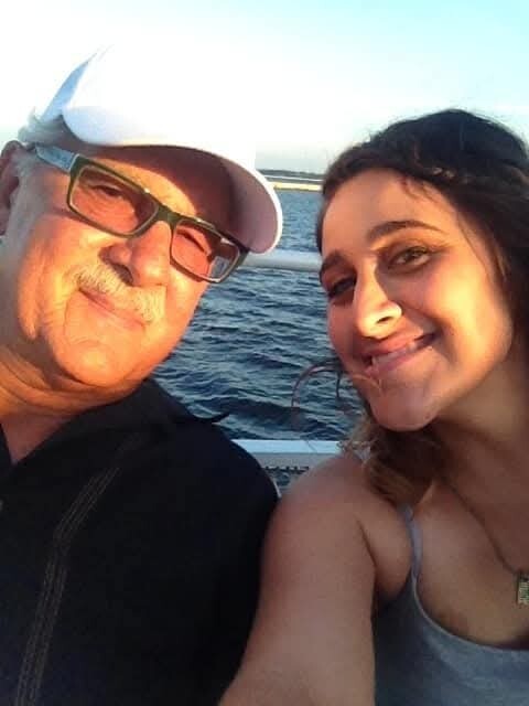 Smiling older man in white cap and glasses with a younger woman on a boat, water in the background (selfie).