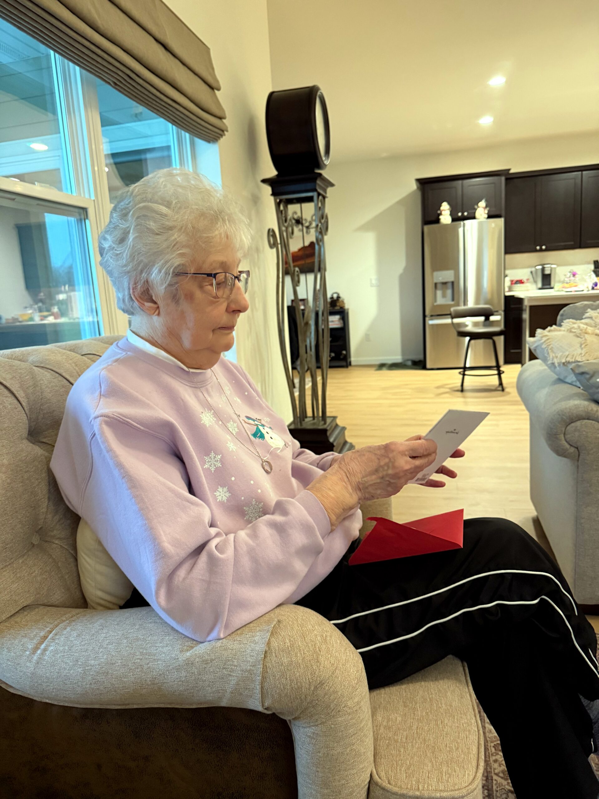 Older woman with white hair and glasses sits on a beige sofa, reading a card in a bright living room.