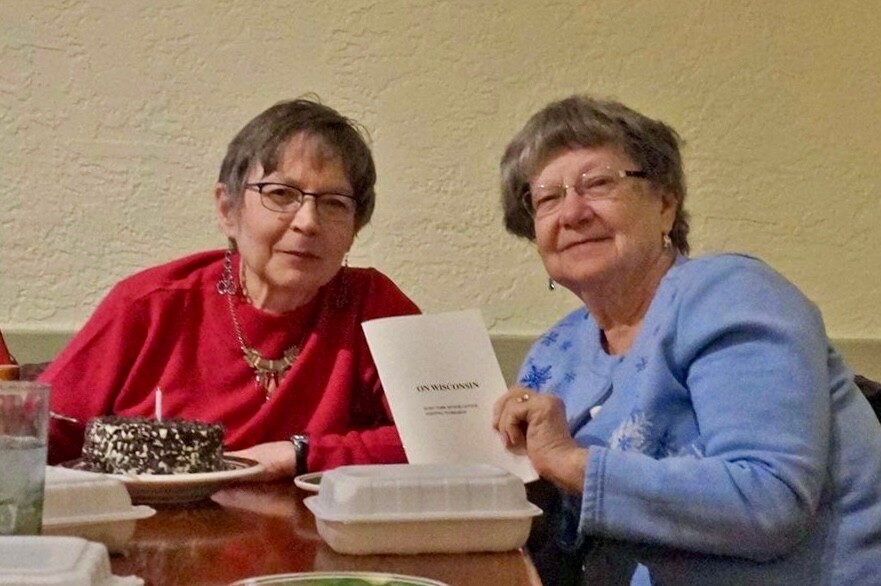 Two elderly women sit at a dining table with a chocolate cake, drinks, and takeout boxes; one holds a card that reads Wisconsin.