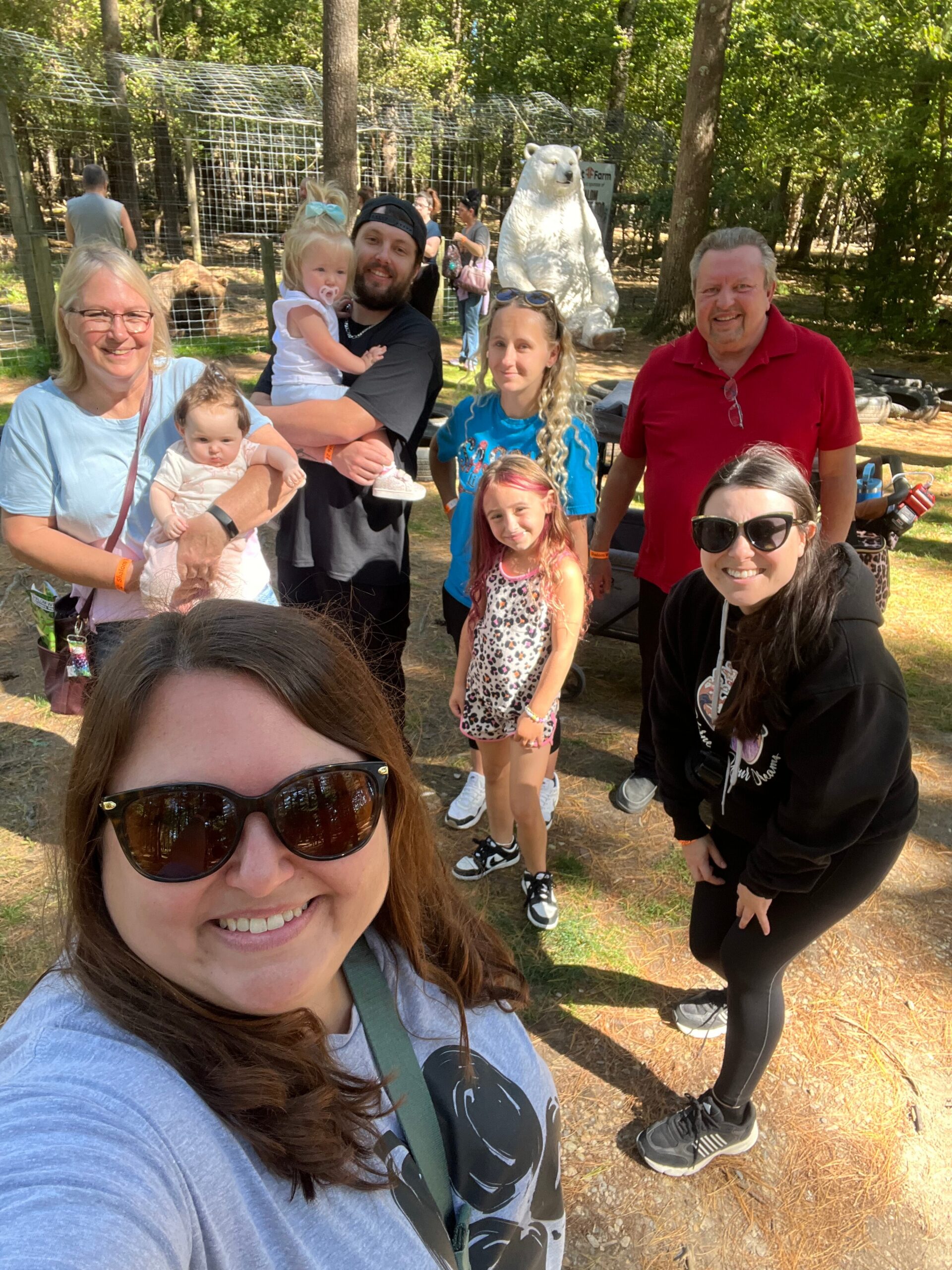 Smiling woman in foreground takes a selfie with a group of people outdoors, including children, near a white polar bear statue in a wooded area behind a fence.