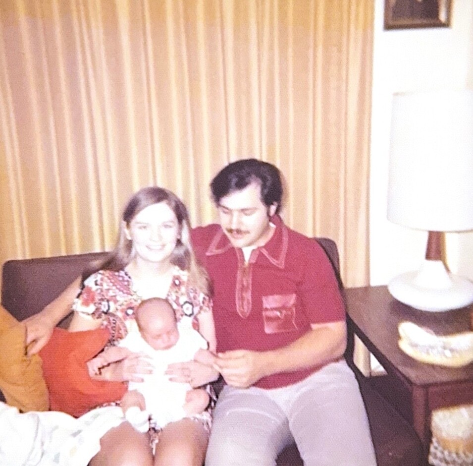 A couple sits on a sofa with their newborn baby between them, smiling at the camera in a living room with striped curtains and a lamp nearby.