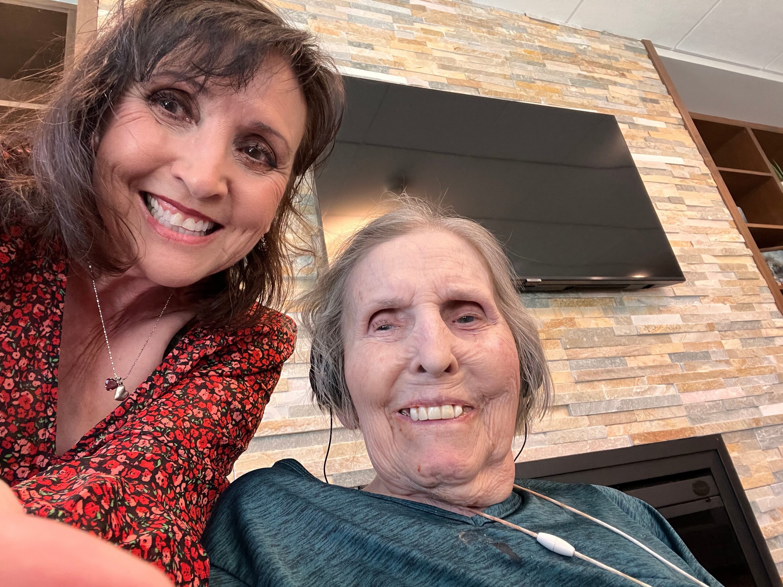 Two smiling women posing for a selfie outdoors, with a stone wall and mounted TV behind them.