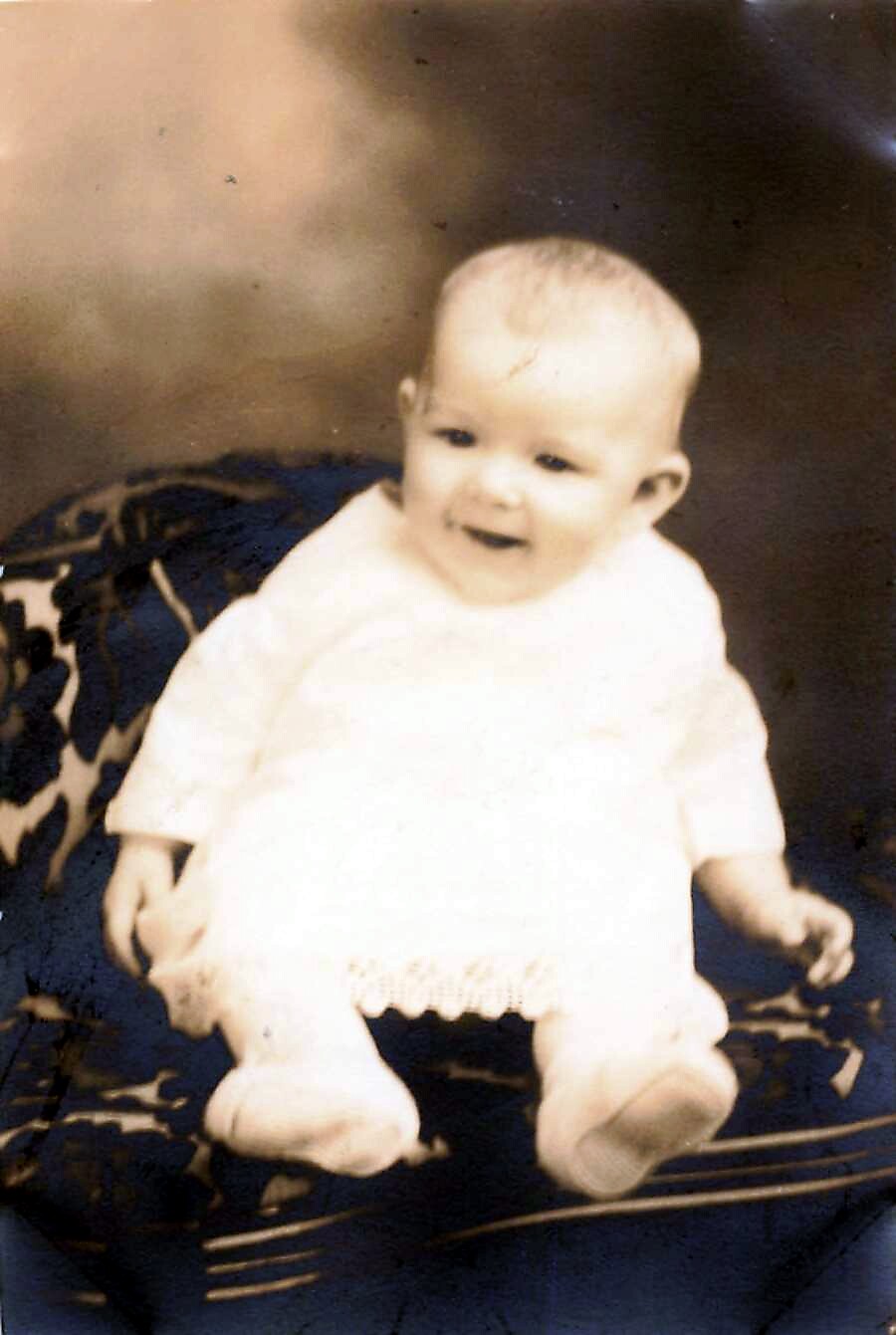 Smiling infant in a white outfit sitting on a dark blue patterned surface.