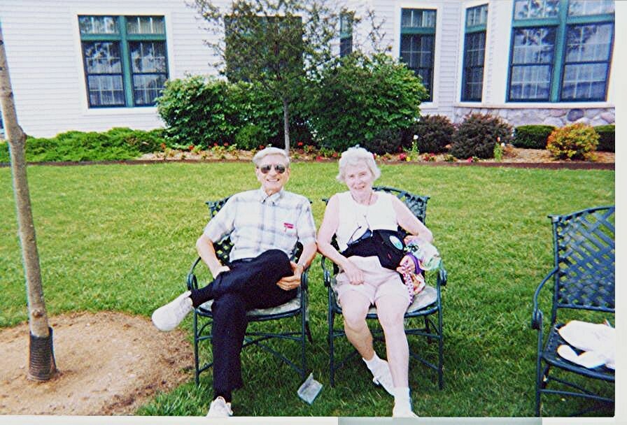 Elderly man and woman sit in metal lawn chairs on a green lawn in front of a white building, smiling at the camera; a baby in a carrier rests on the woman's chest.
