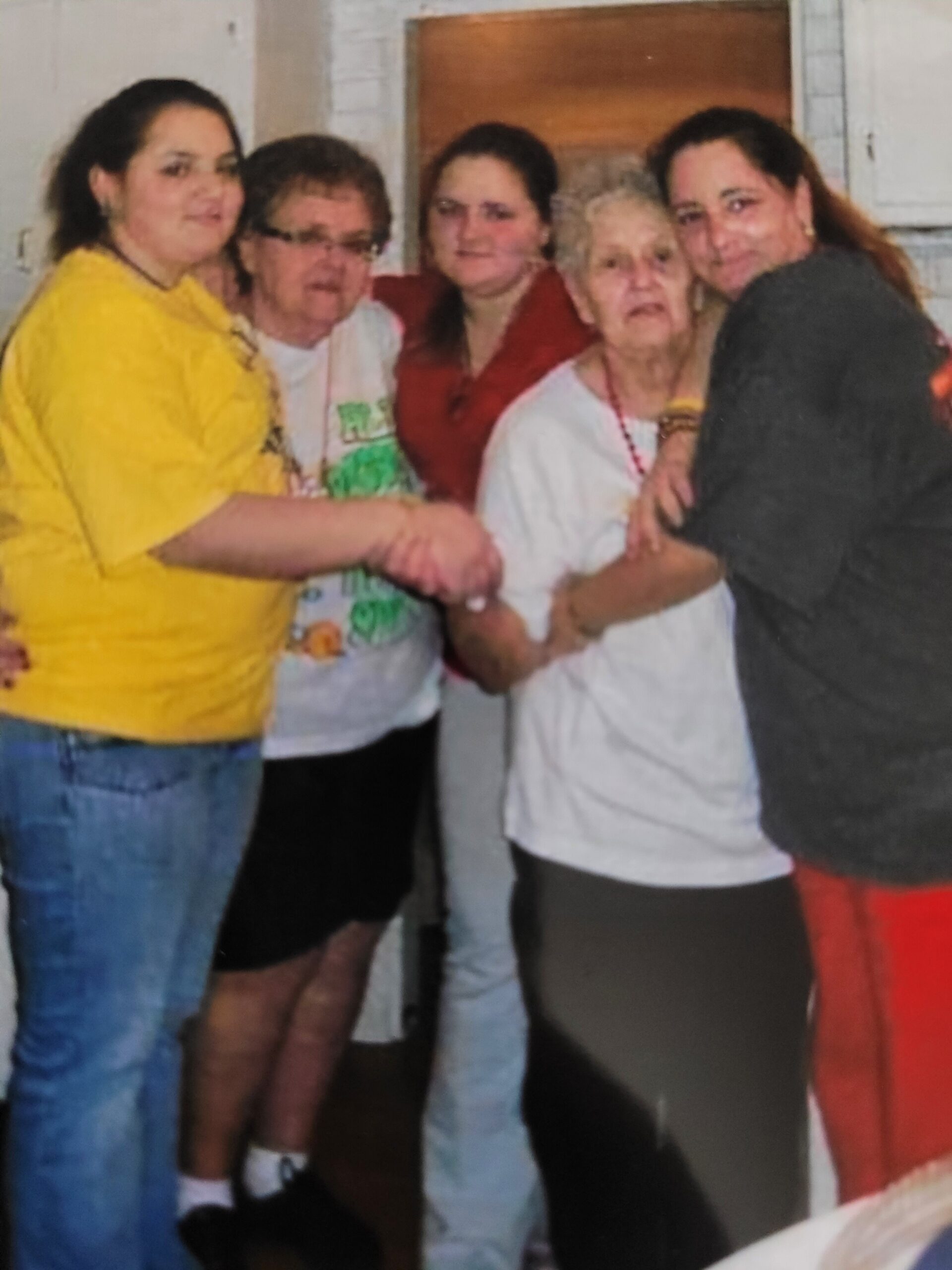 Group of five women of varying ages standing close together in a kitchen, arms linked and smiling at the camera.