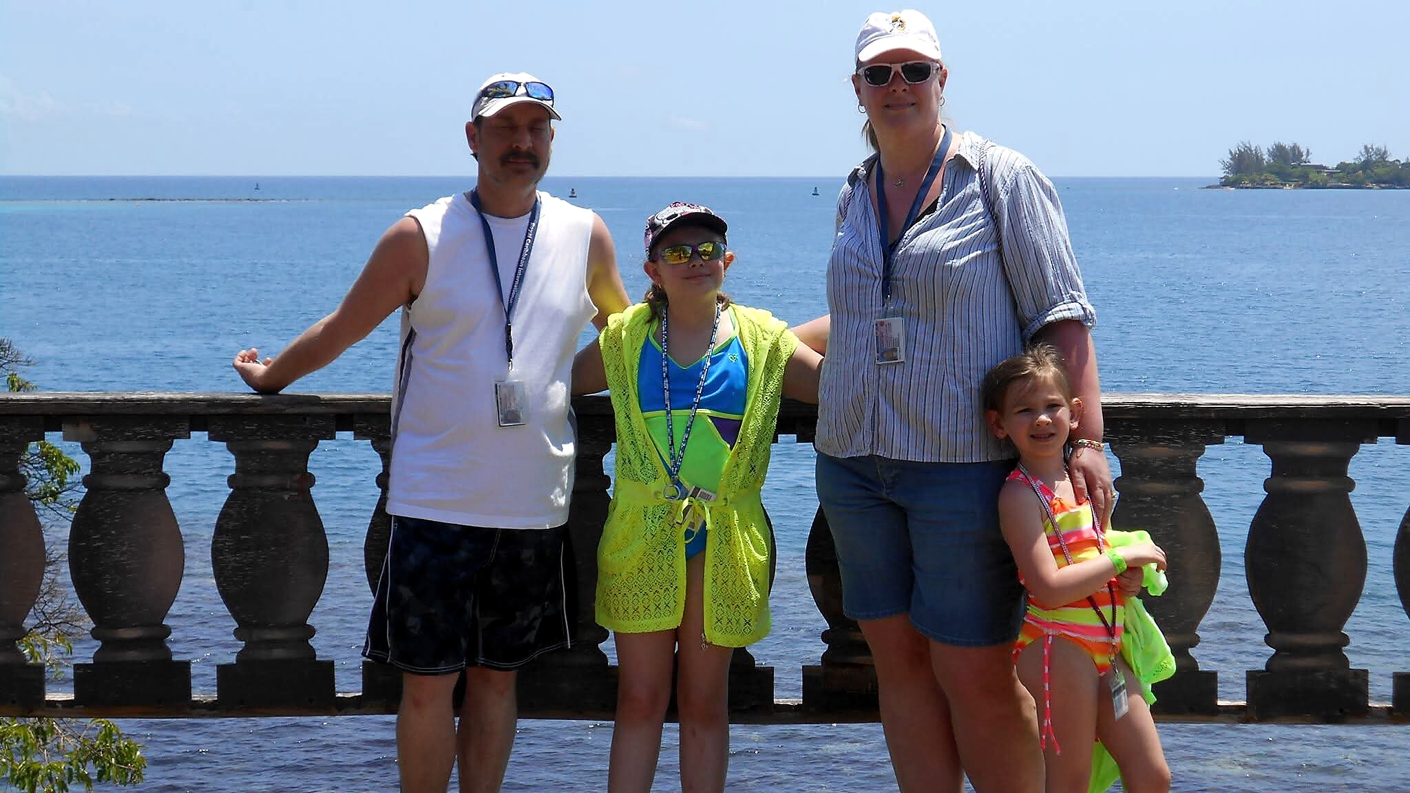 Family of four posing by a seaside railing with a blue ocean backdrop on a sunny day, wearing bright summer clothes and badges around their necks.