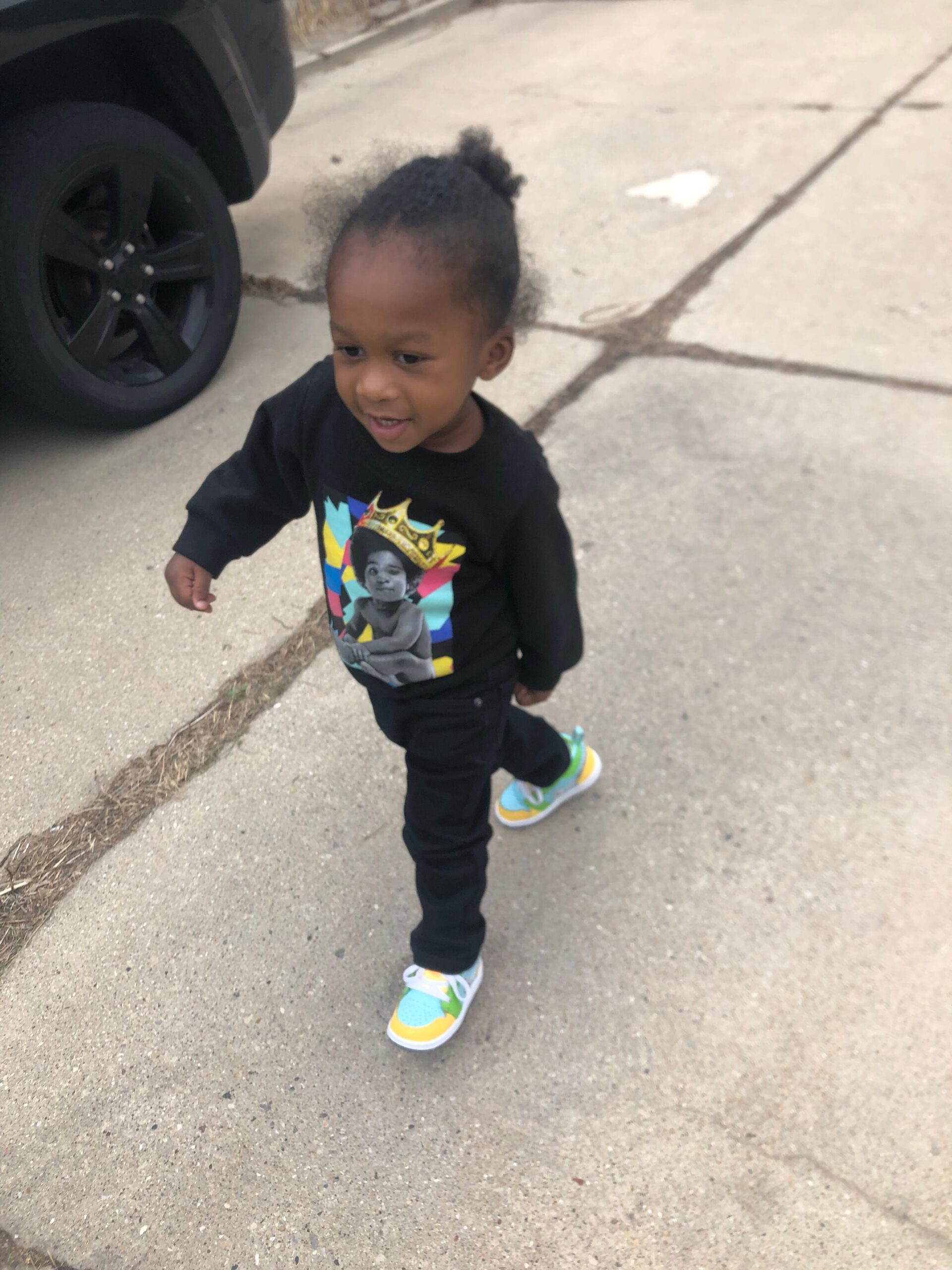 Young child walking on a sidewalk near a black SUV tire, wearing a black shirt with a colorful graphic and bright sneakers.