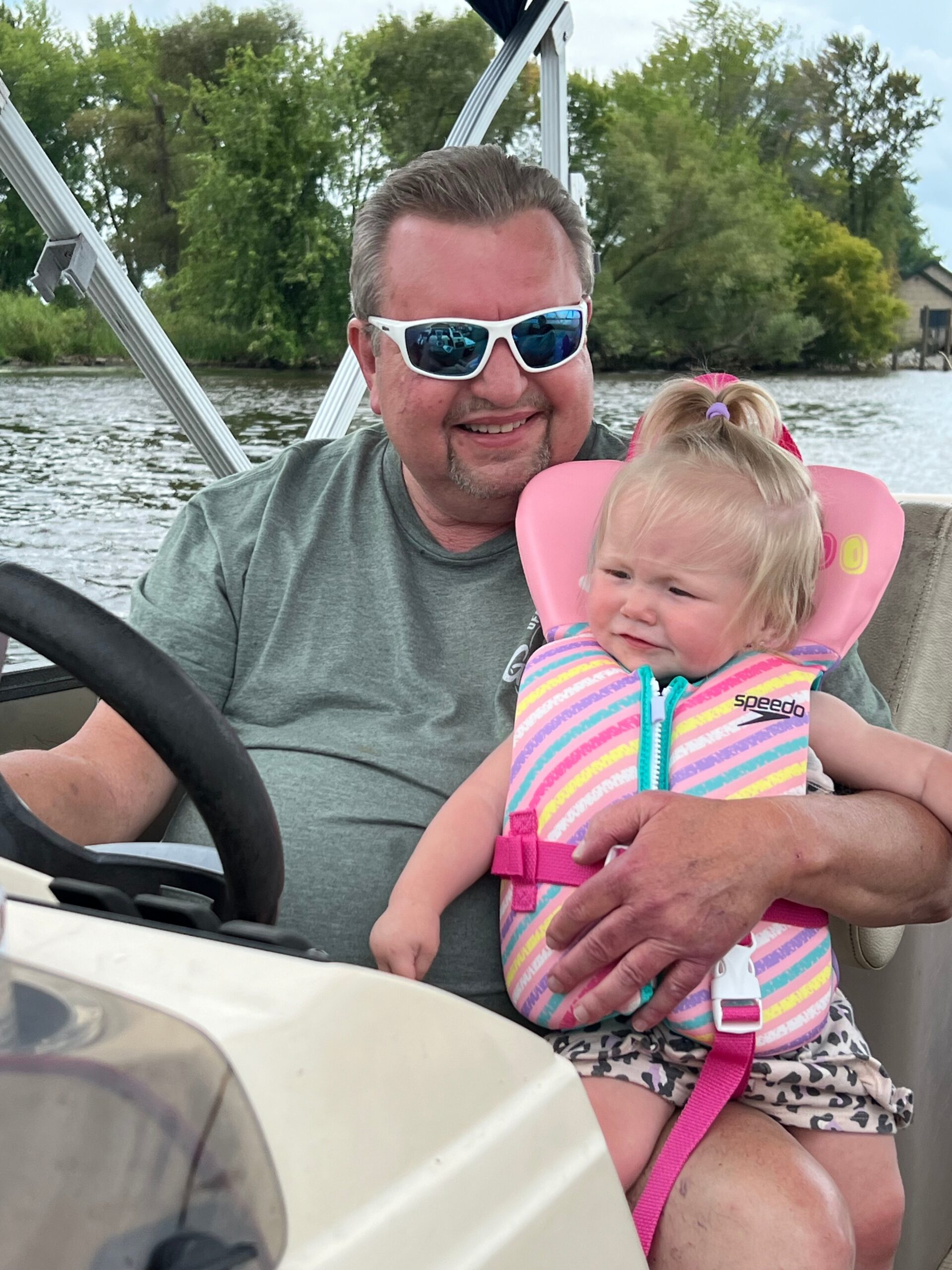 Man wearing white sunglasses smiles while steering a boat with a toddler in a pink life jacket beside him, water and trees in the background.