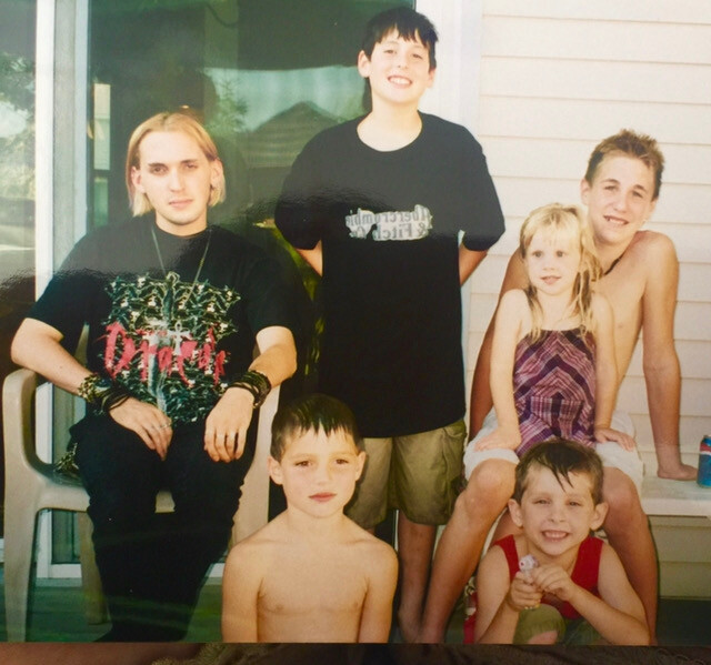 Six siblings posing on a porch: two shirtless boys in front, a girl in a purple dress seated with a smiling teen, and three teens behind them.