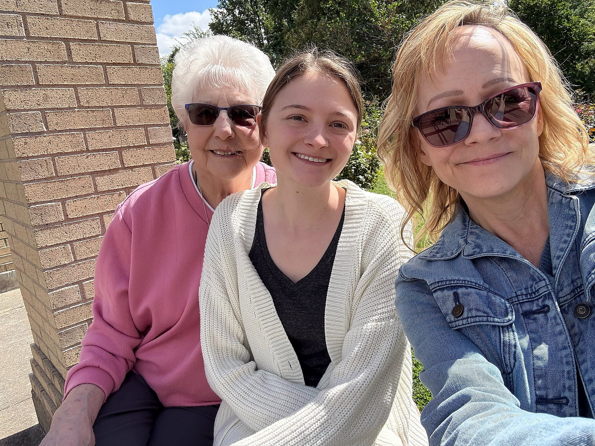 Three women posing for a selfie outdoors beside a brick wall, smiling with greenery in the background.