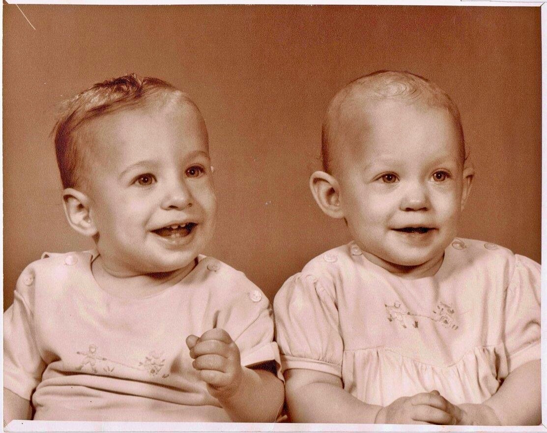 Two smiling toddlers sit side by side in matching light outfits against a warm brown studio backdrop.