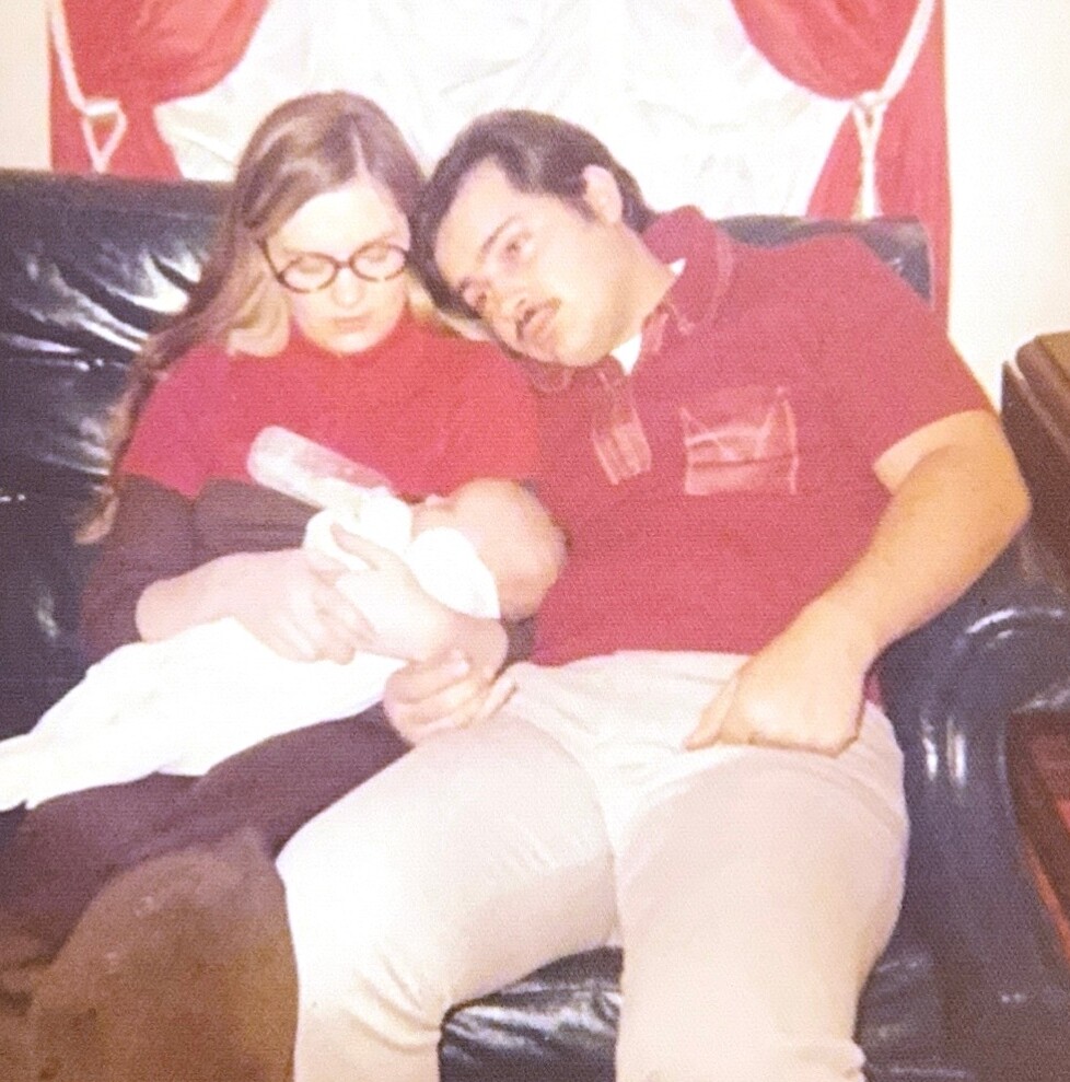 Young couple cuddling on a black leather couch with their newborn baby wrapped in white. The mother holds the baby while the father leans in close.