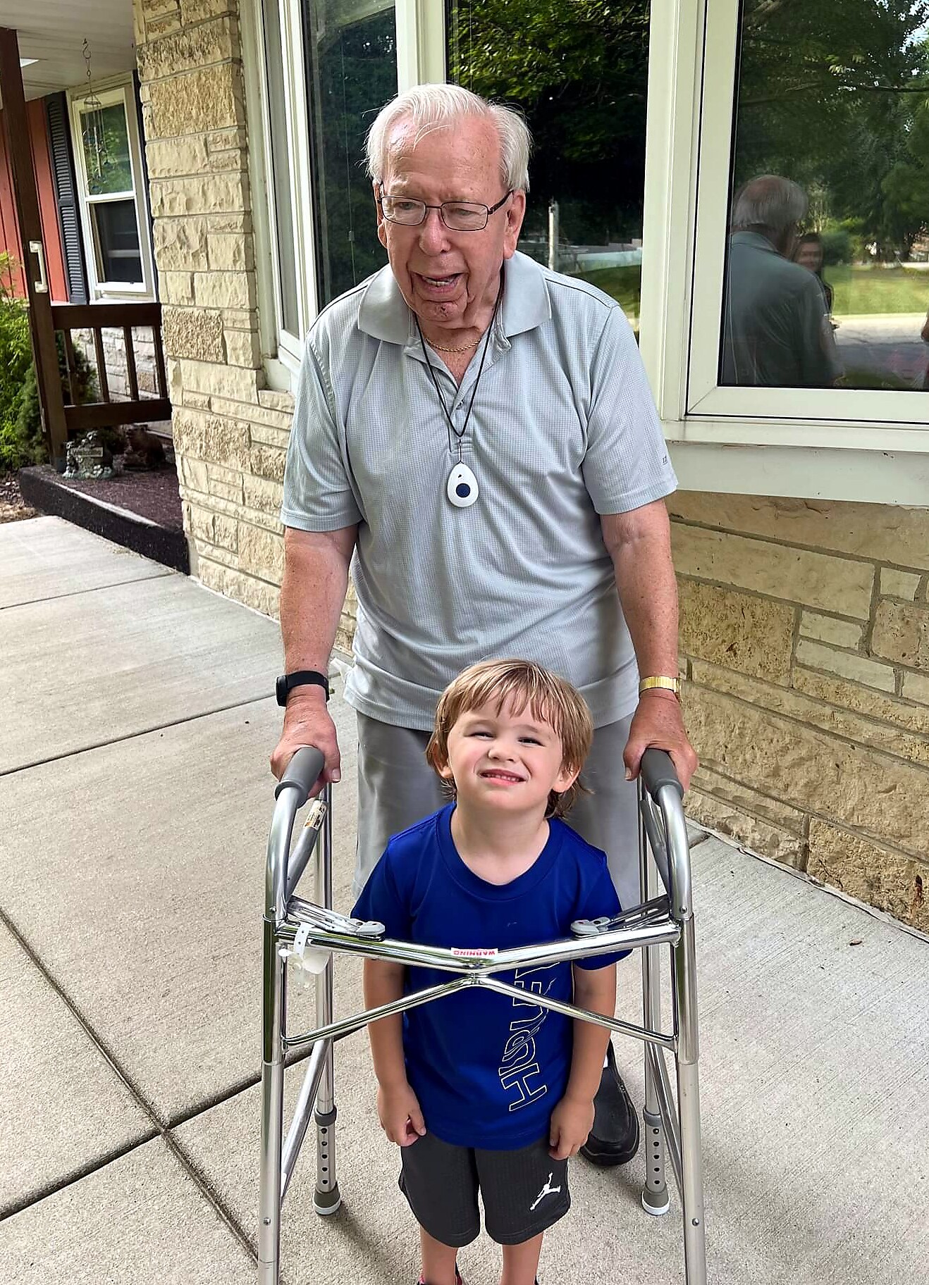 Older man with white hair and glasses pushing a walker beside a smiling young boy outside a brick house with a window nearby.