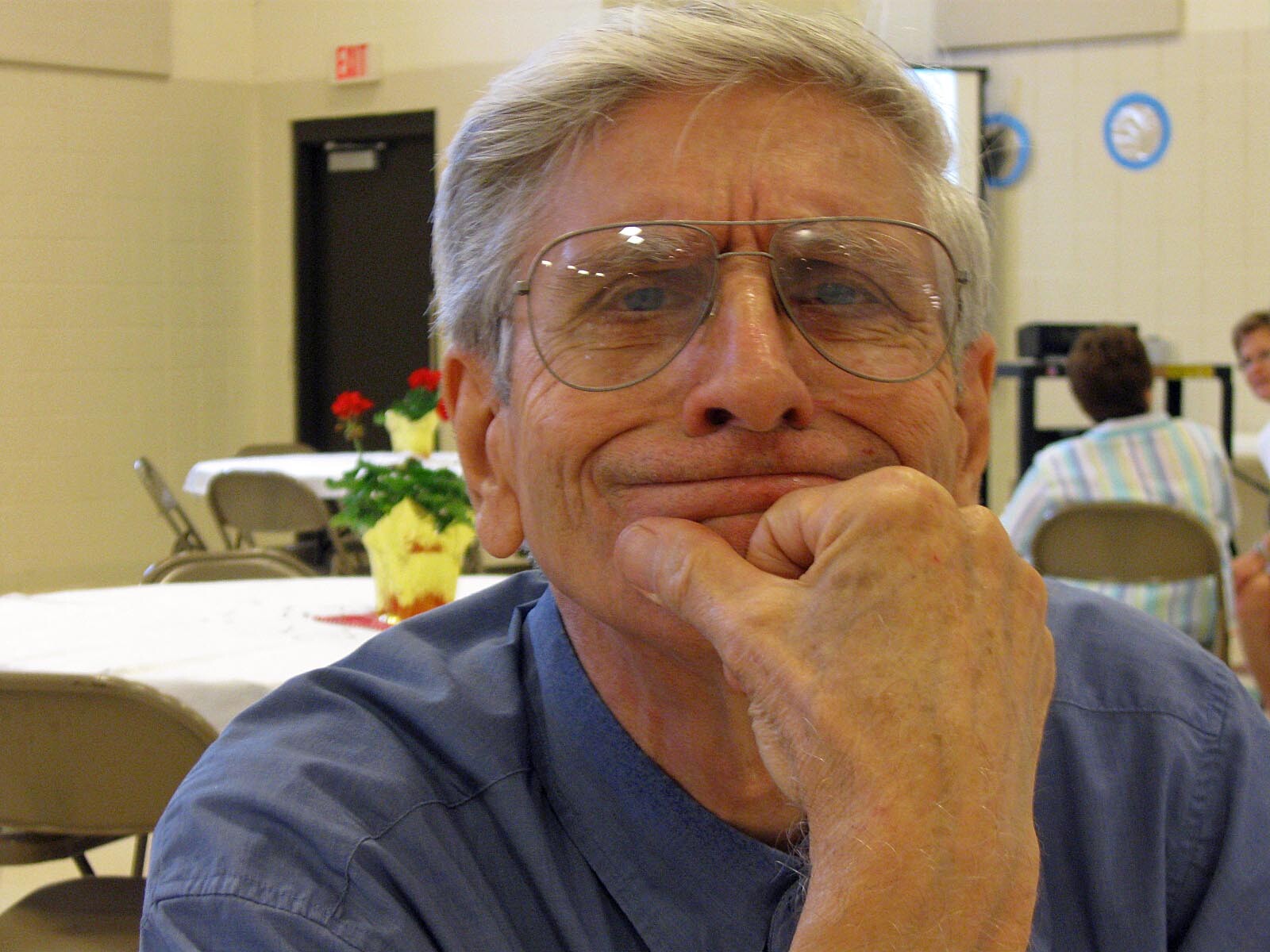 Close-up of an older man with gray hair and large glasses resting his chin on his hand at a table.
